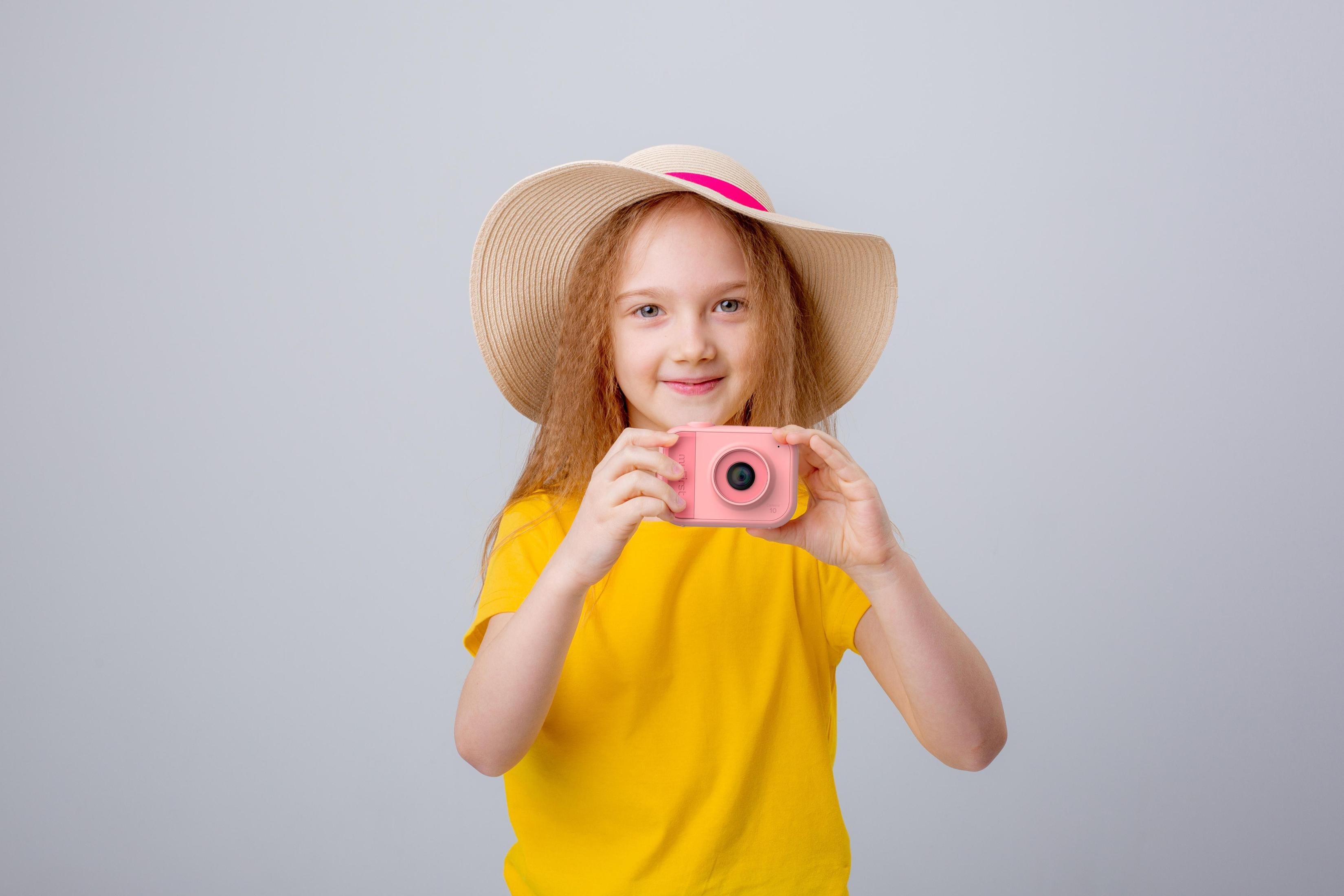 Een jong meisje in een geel shirt en hoed houdt een roze camera vast en glimlacht naar de kijker.