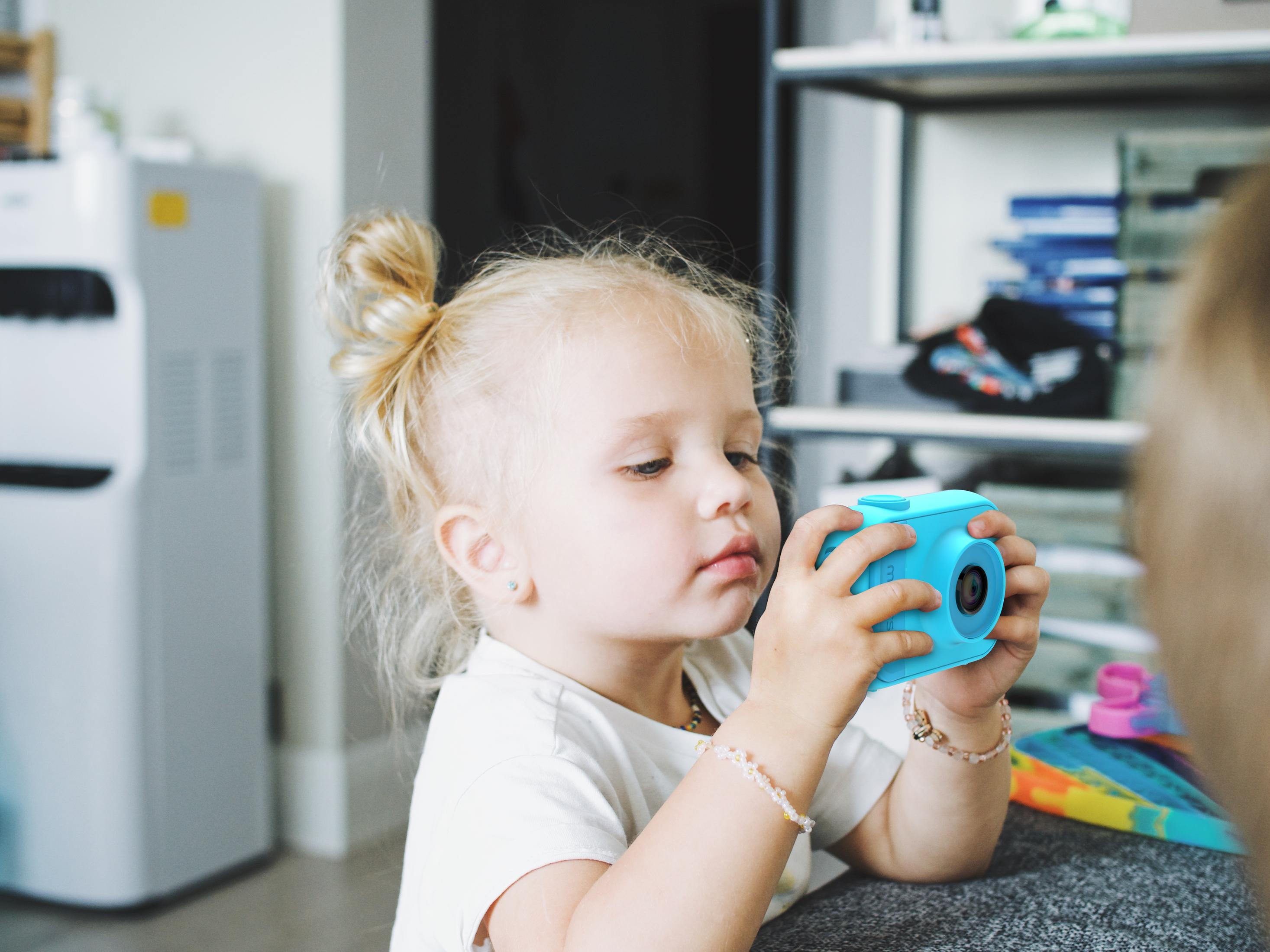 Ein junges Mädchen hält drinnen eine blaue Spielzeugkamera. Sie hat blondes Haar und ein weißes Oberteil.