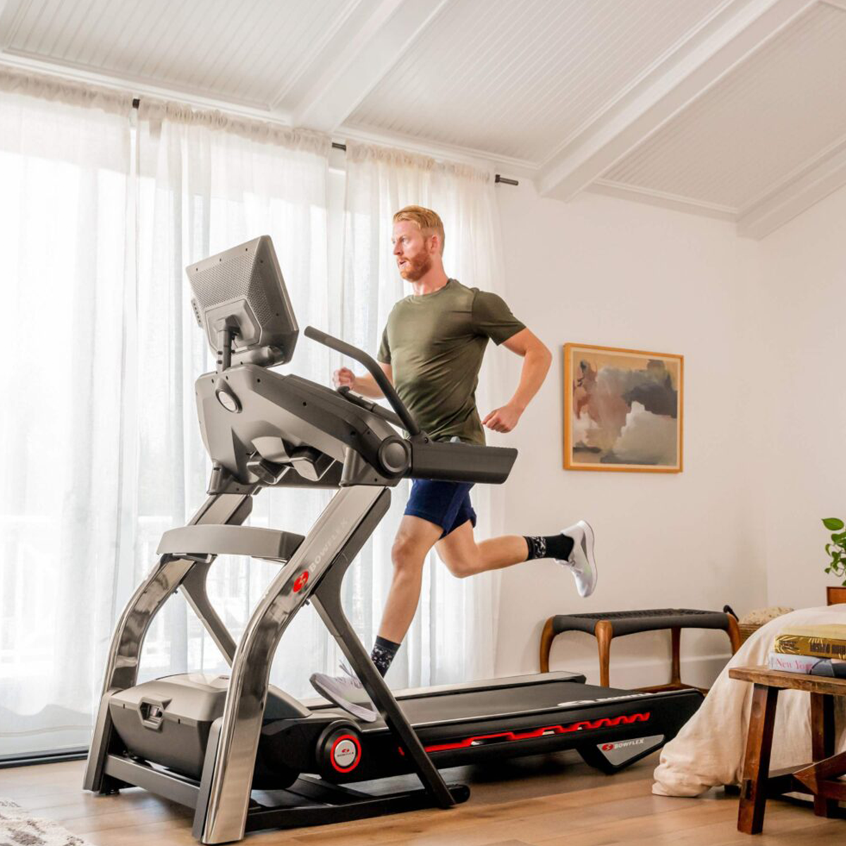 Ein Mann läuft auf einem Laufband in einem Raum mit Fenster und Gemälde. Das Laufband hat einen Bildschirm.