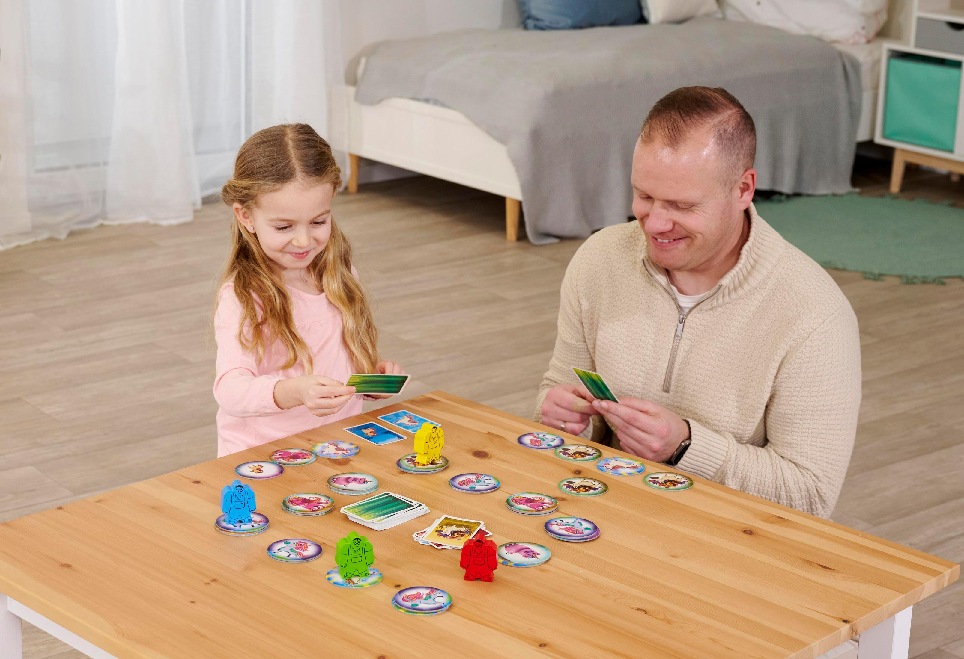 Ein Mädchen und ein Mann spielen ein Brettspiel auf einem Holztisch, umgeben von Spielsteinen. Im Hintergrund ist ein Bett.