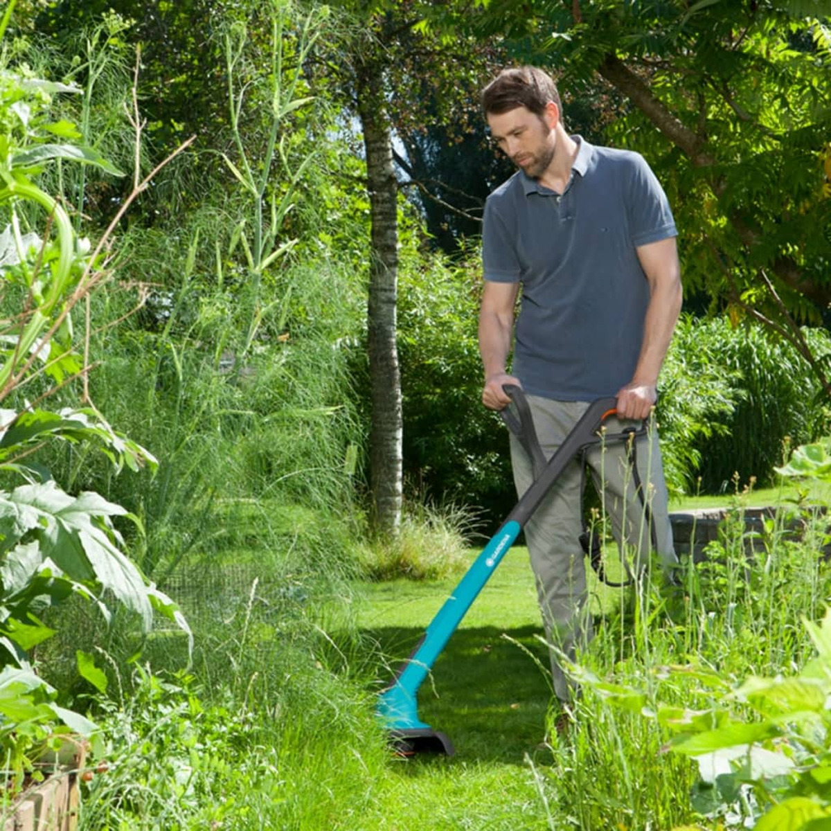 Ein Mann schneidet Gras mit einem teal-schwarzen Rasentrimmer in einem grünen Garten. Üppiges Grün umgibt ihn.