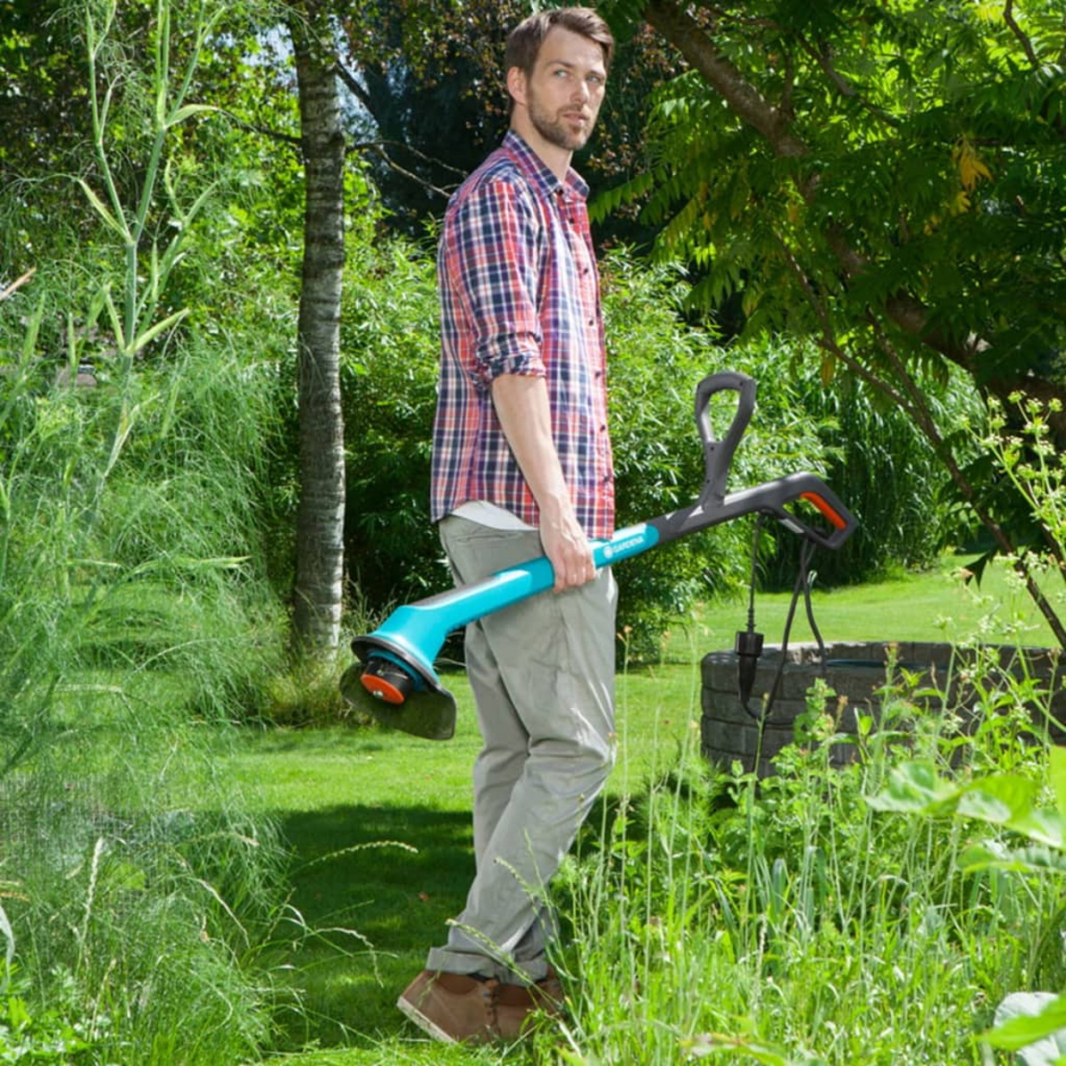 Ein Mann in kariertem Hemd steht in einem Garten und hält einen blau-schwarzen Rasentrimmer.