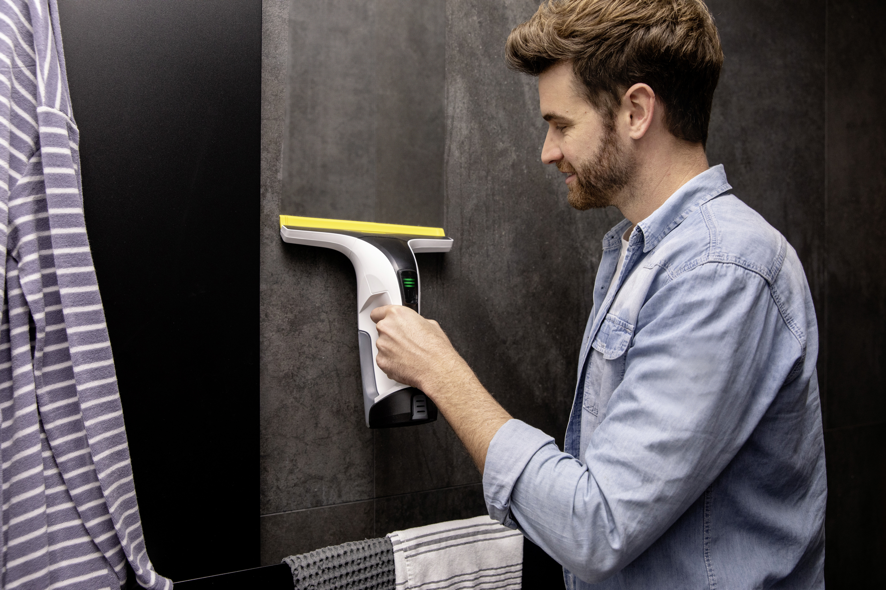 Mężczyzna używa myjki do okien na czarnej kafelkowej ścianie, z ręcznikiem i szlafrokiem.