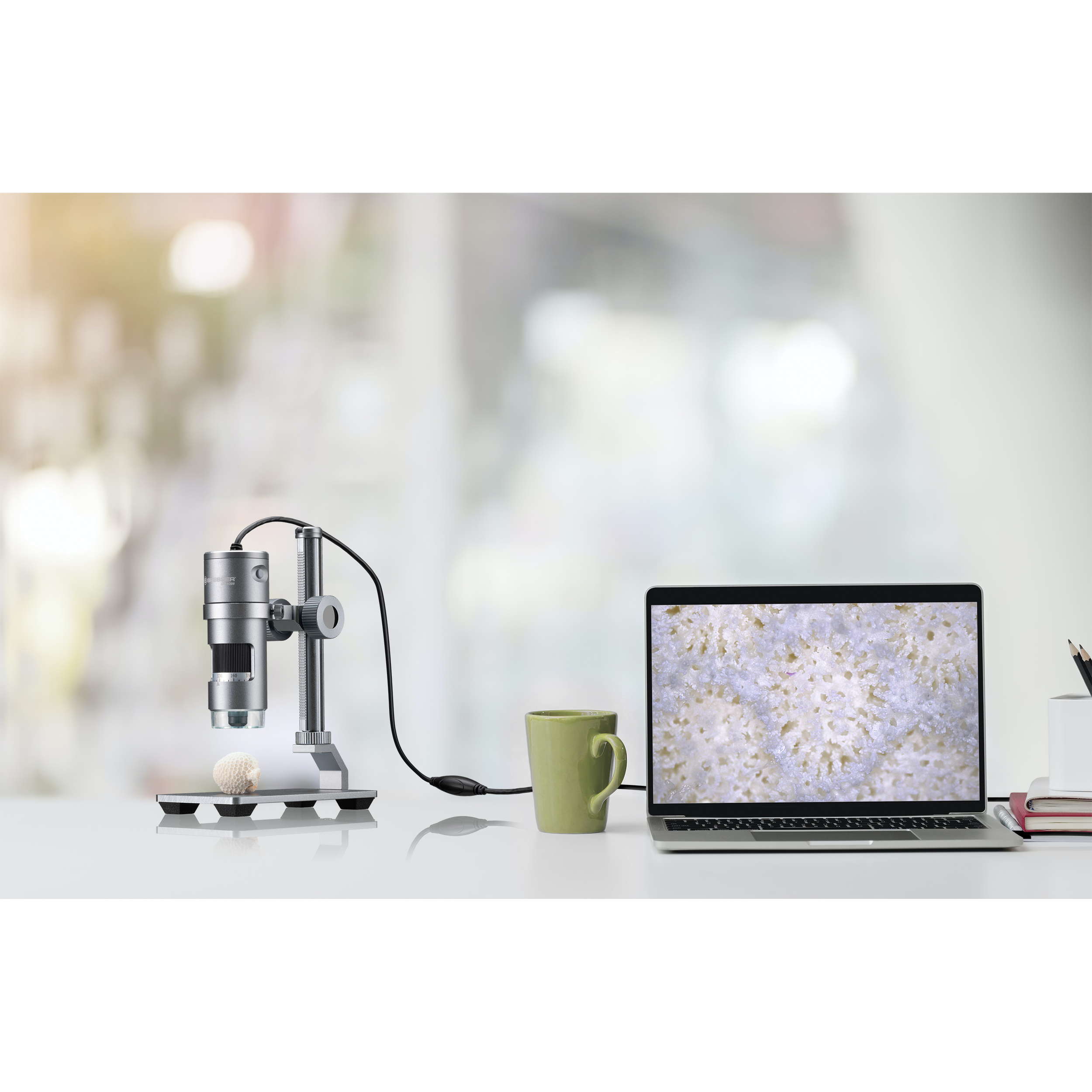 Ein Mikroskop mit einem Laptop, der eine Probe zeigt, eine Tasse und ein verschwommener Hintergrund eines Arbeitsplatzes.