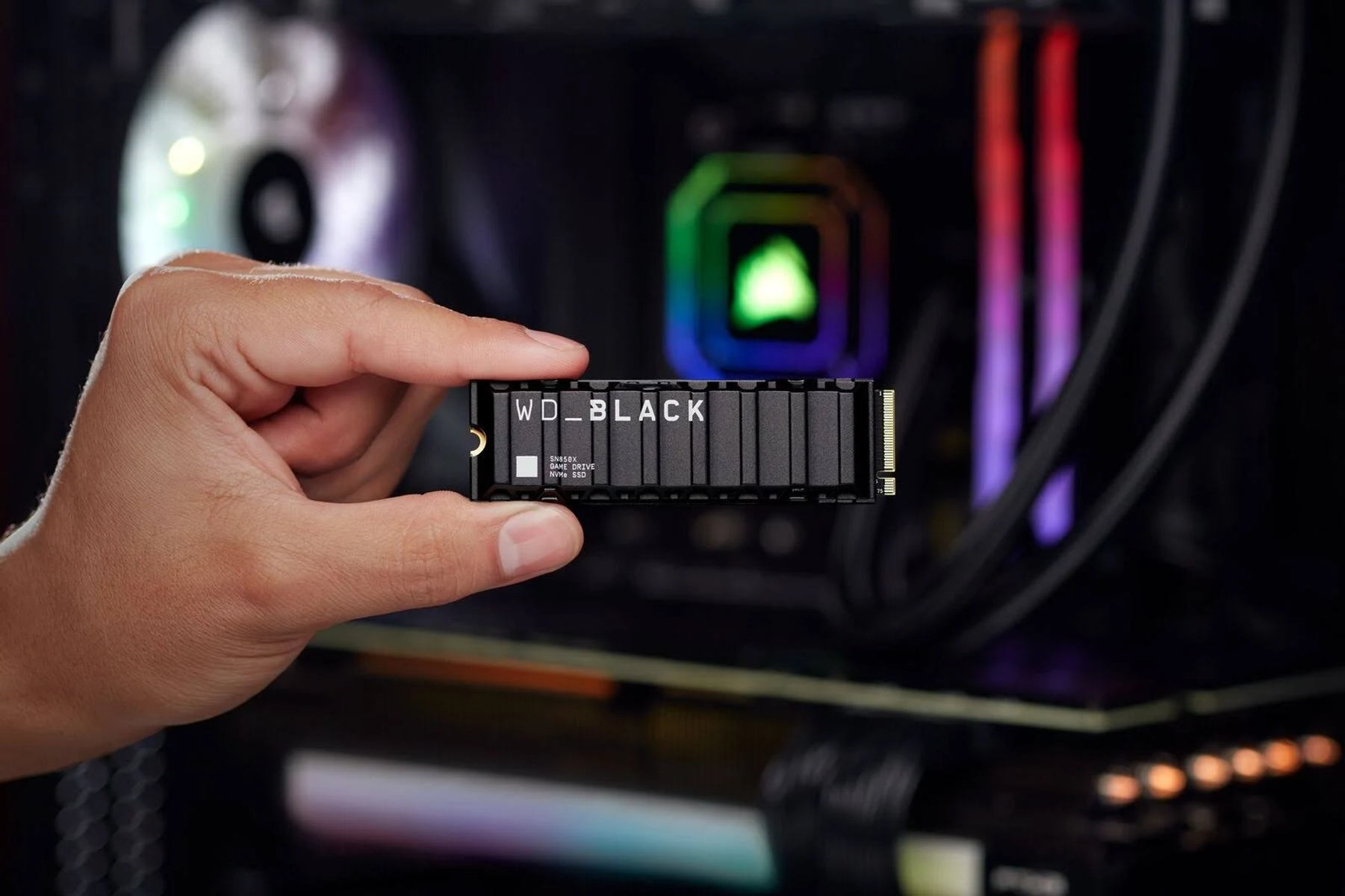 Dłoń trzymająca czarny dysk SSD z białym tekstem, w obudowie komputera ze światłami.