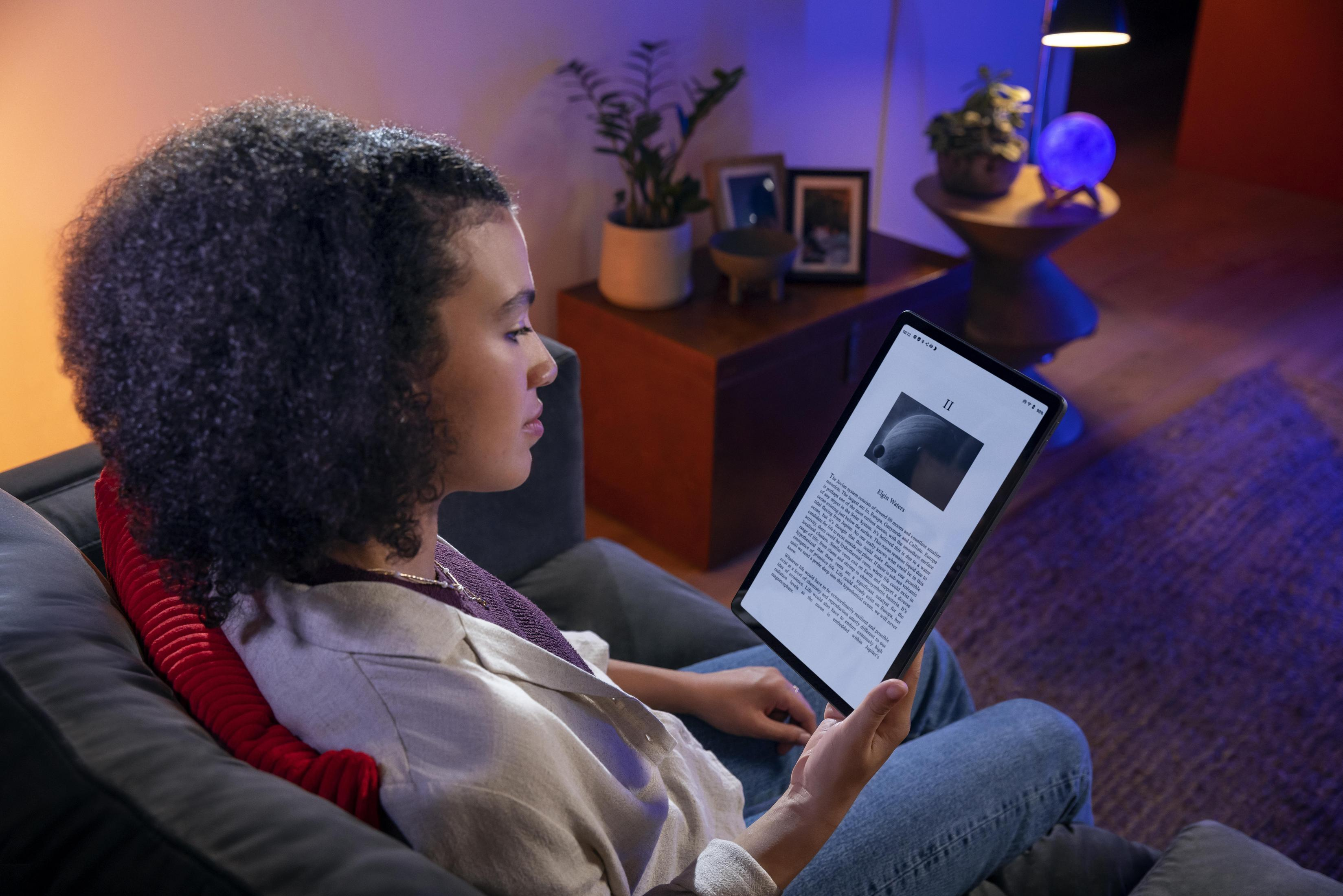 Frau liest Tablet in schwach beleuchtetem Raum, auf Couch sitzend. Pflanze und Lampe im Hintergrund.
