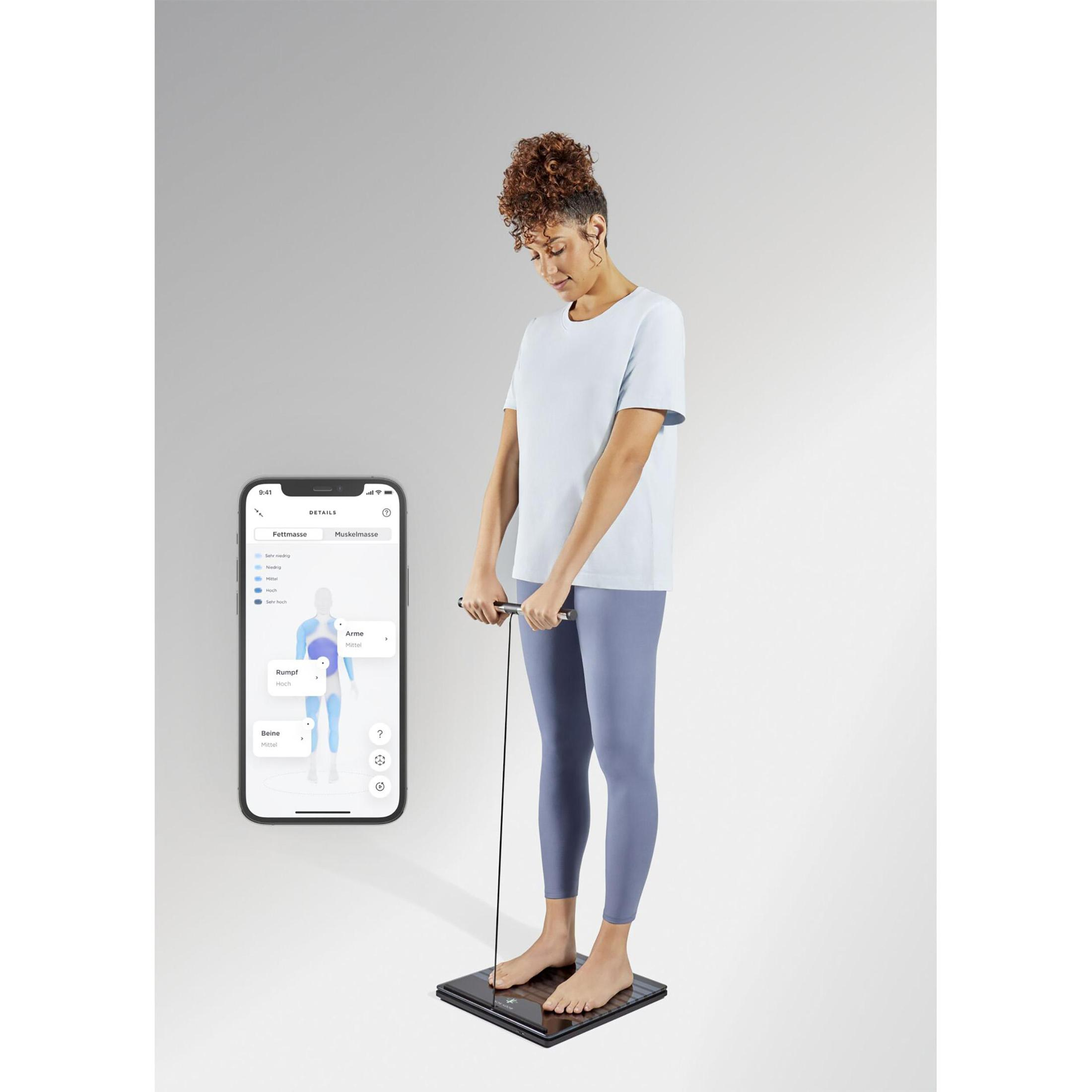 Kobieta na wadze trzymająca urządzenie pomiarowe, przegląda dane składu ciała w telefonie.