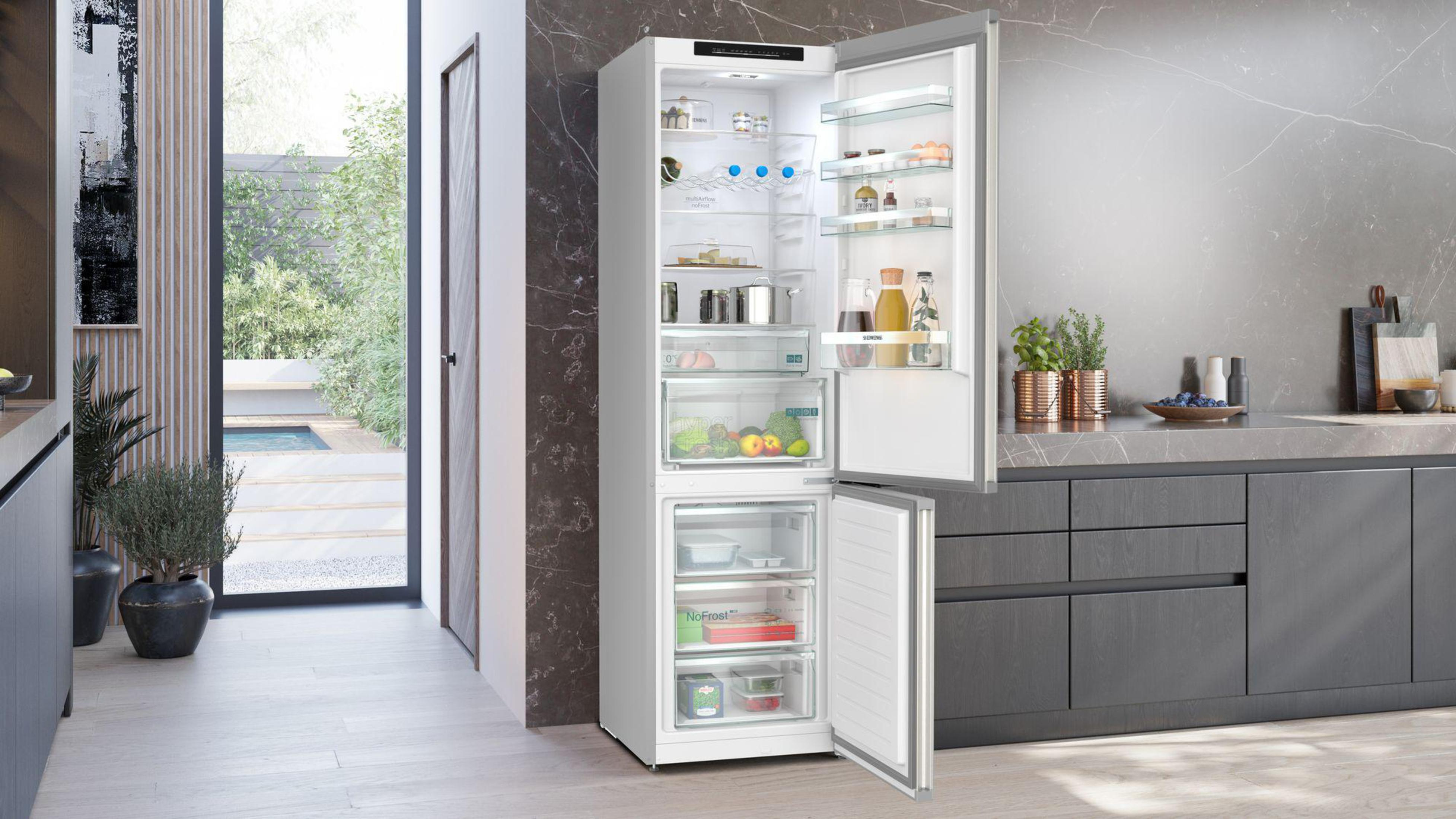 Un refrigerador de acero inoxidable está abierto, mostrando estantes con comida, verduras y bebidas. Una cocina está en el fondo.