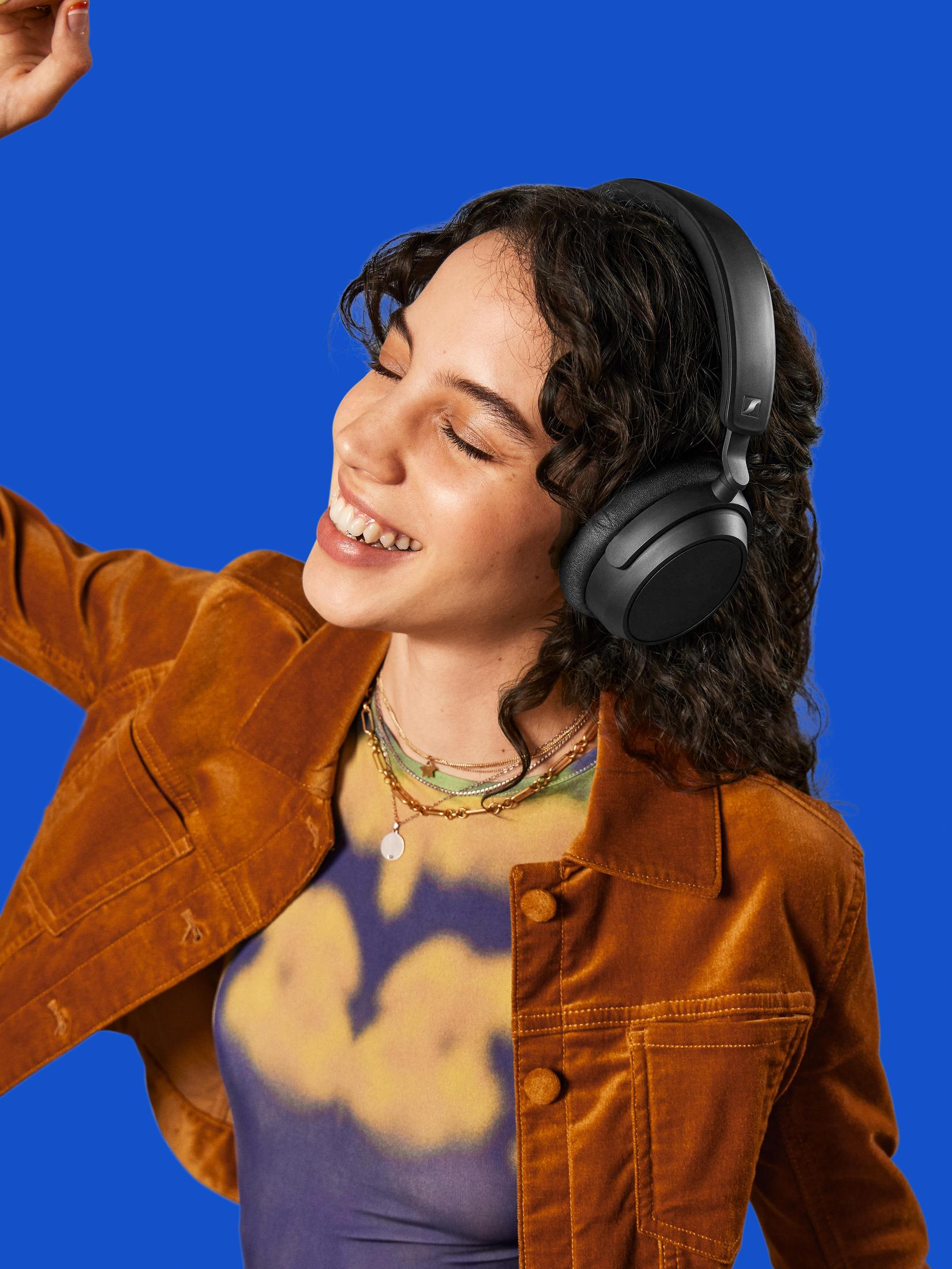 Donna con cuffie sorridente su sfondo blu. Indossa giacca marrone e gioielli.