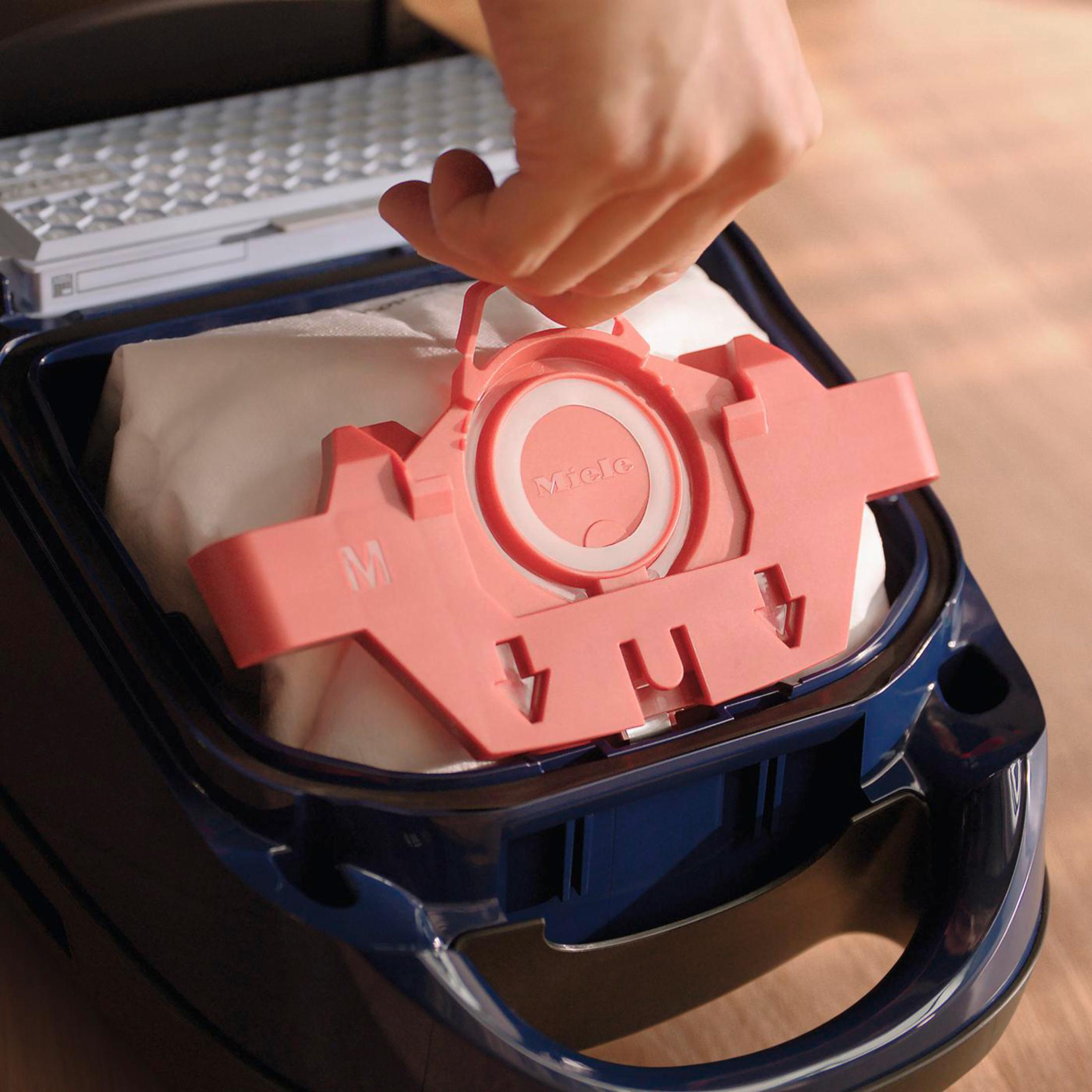 Eine Hand entfernt einen rosa Filterhalter aus einem dunkelblauen Staubsaugerbeutel-Fach.