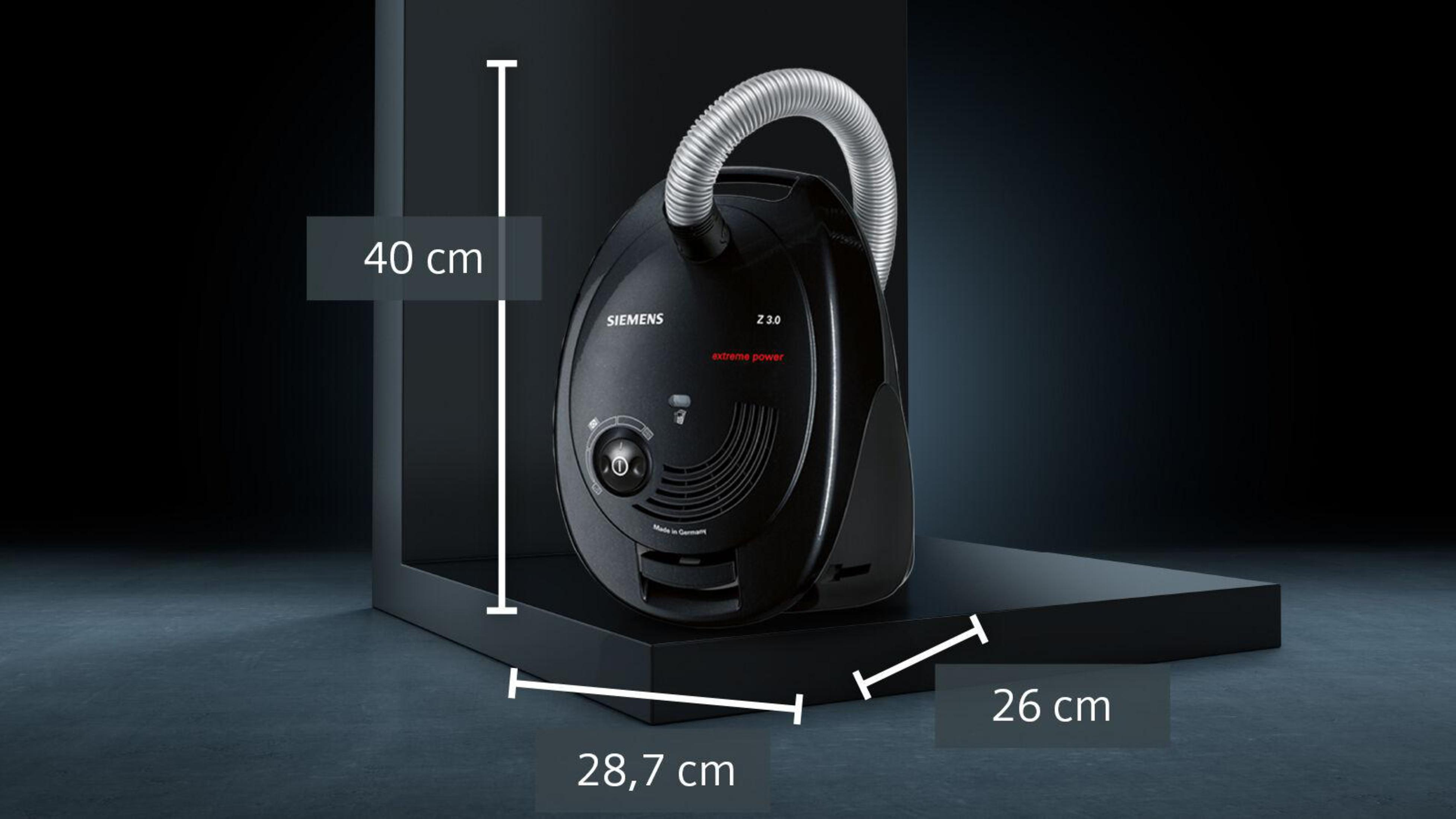 Czarny odkurzacz w czarnym rogu. Podano wymiary wysokości i szerokości.