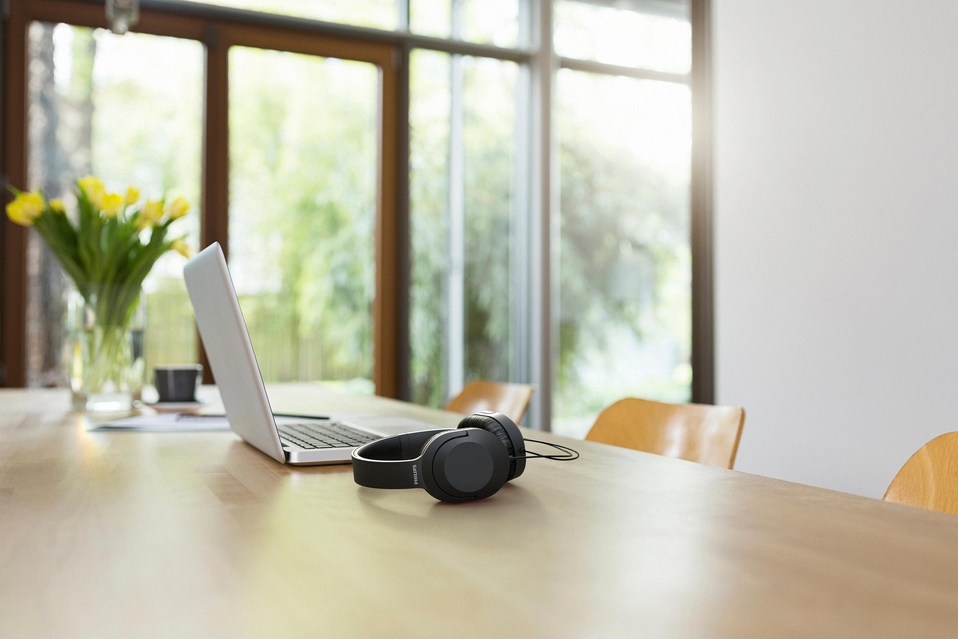 Laptop i czarne słuchawki na drewnianym stole. Wazon z żółtymi kwiatami po lewej.