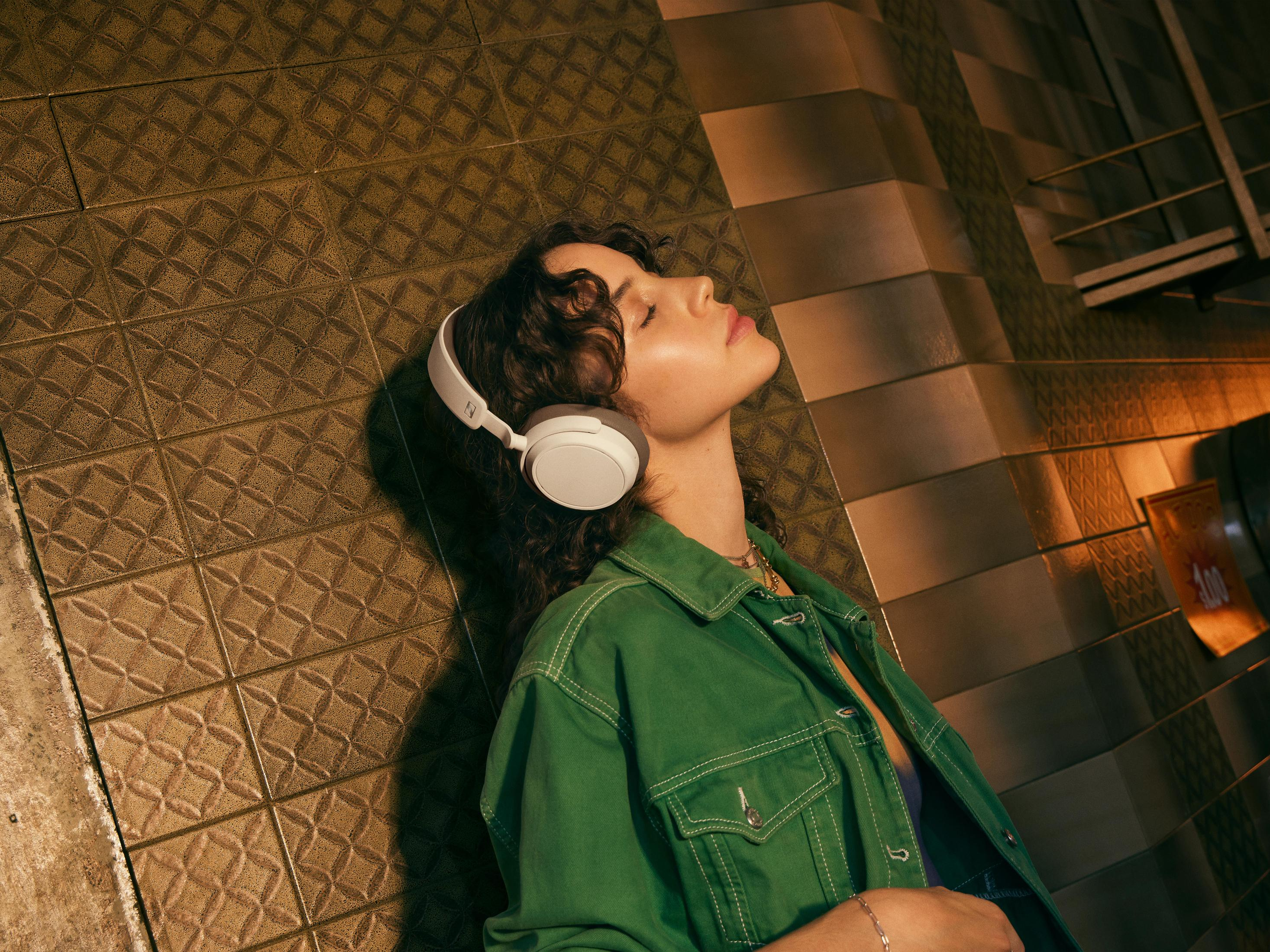 Kobieta z białymi słuchawkami, głową opartą o ścianę. Zielona kurtka, brązowe płytki i jasnobrązowe włosy. Oczy zamknięte.
