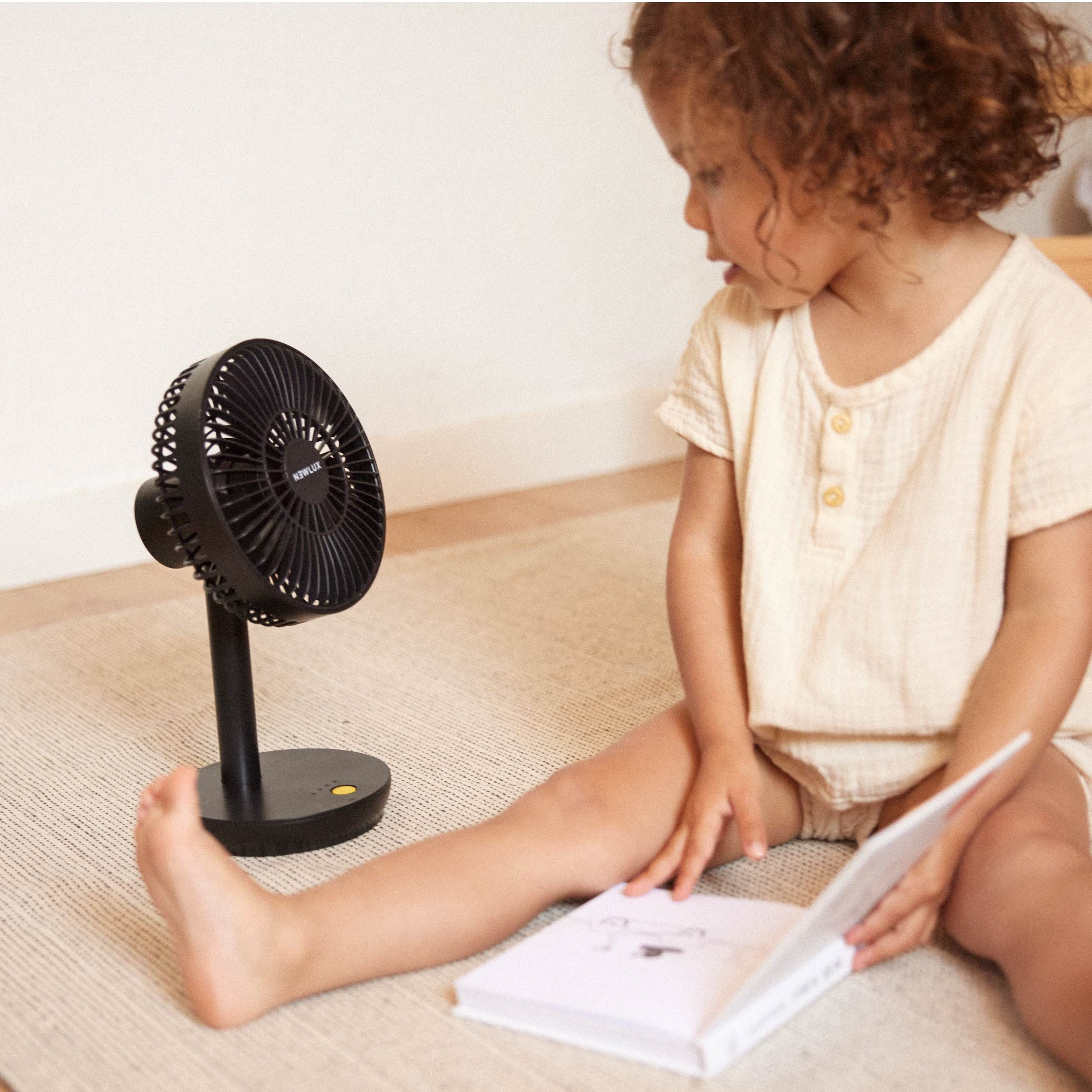 Un niño está leyendo mientras un pequeño ventilador está a su lado. Fondo beige.