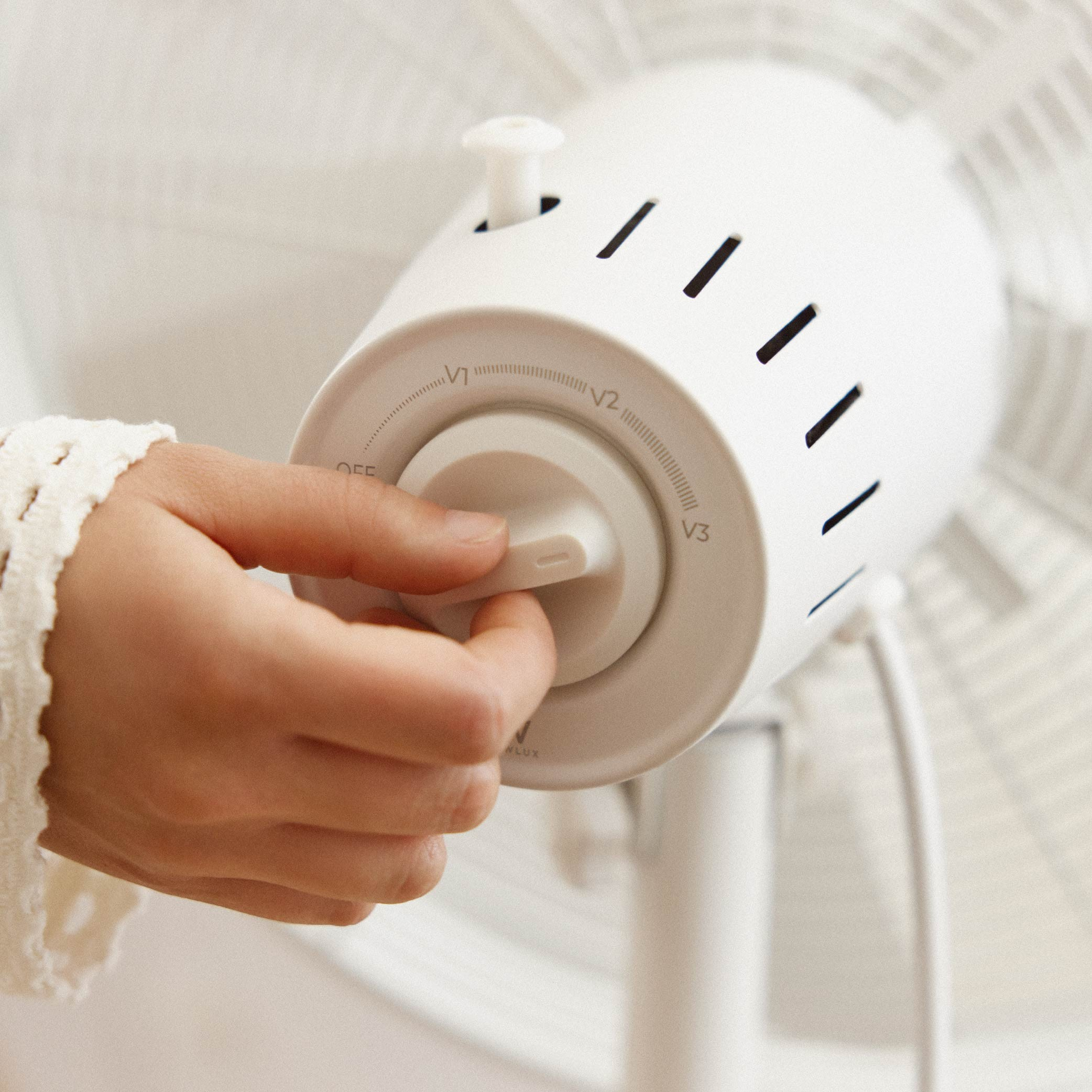 Una mano gira el dial de velocidad de un ventilador blanco, con aspas visibles al fondo.