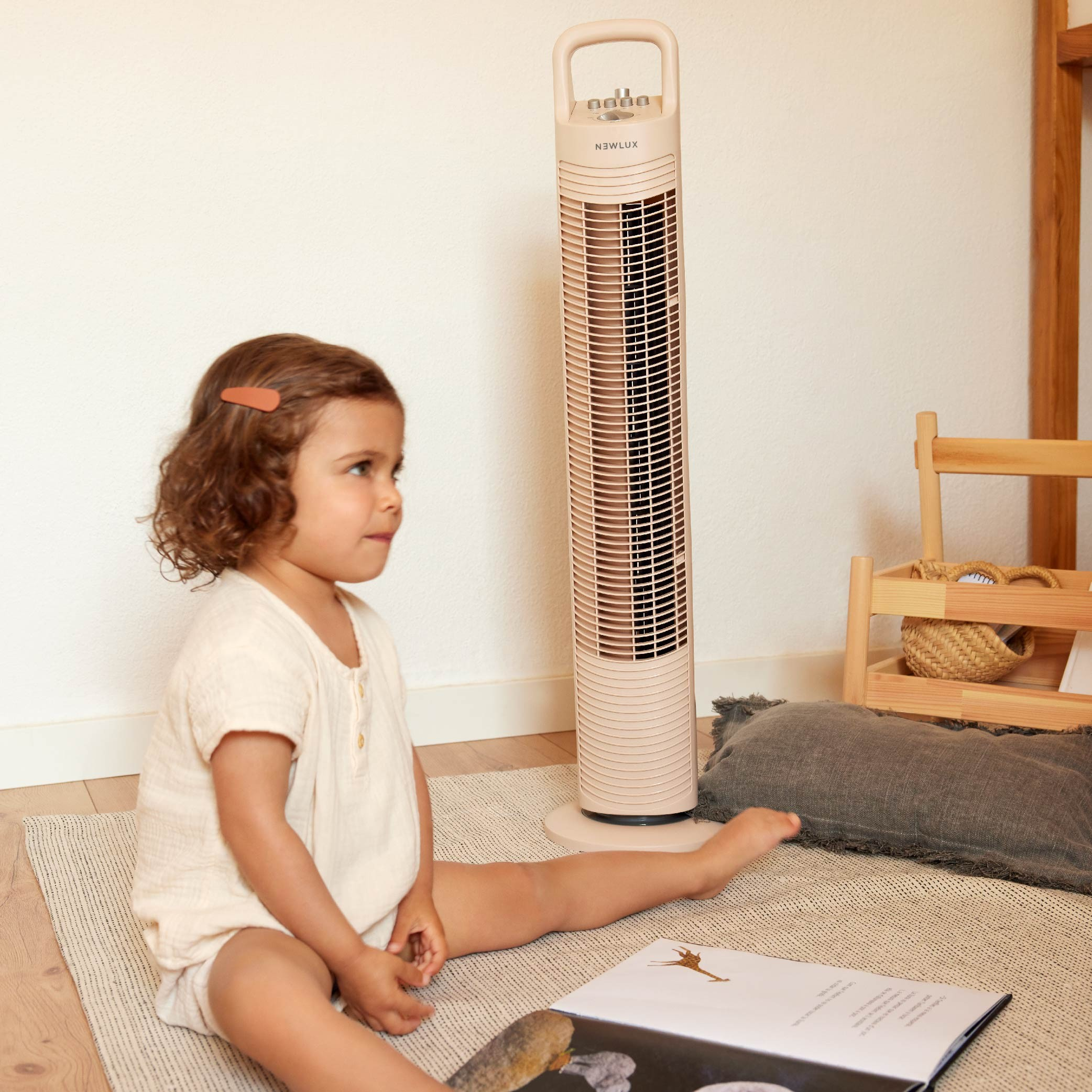 Una niña pequeña se sienta cerca de un ventilador beige. Mira hacia la izquierda. Se ve una alfombra y un libro.