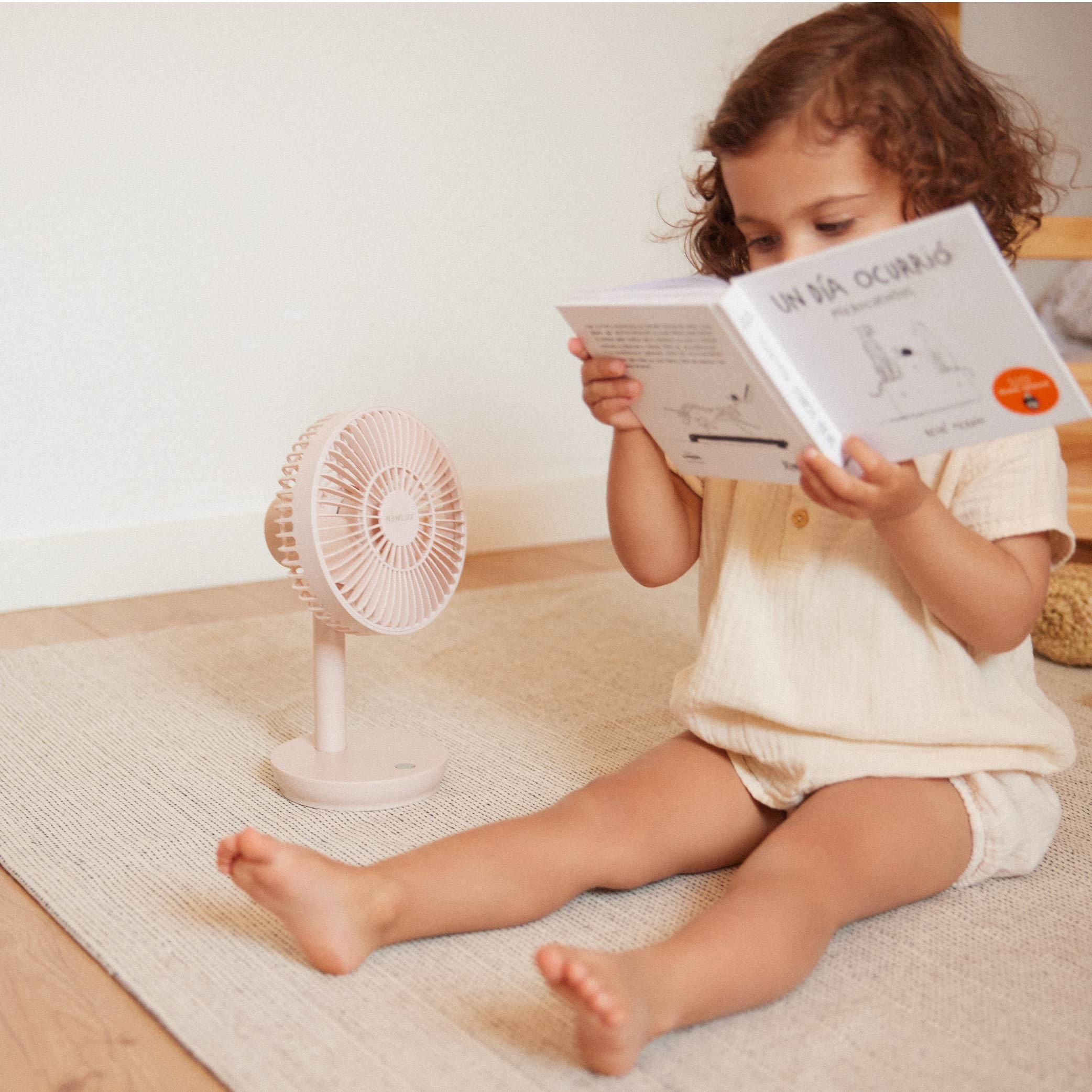Niña leyendo un libro cerca de un ventilador rosa. Está sentada en una alfombra.