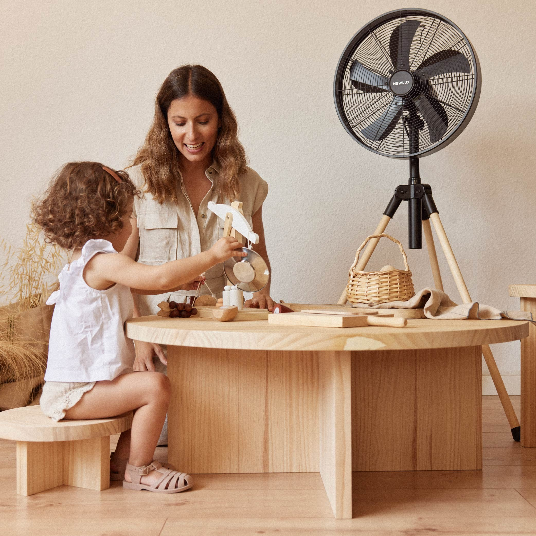 Mujer y niño en la mesa. Ventilador al fondo. Niño juega con juguete. Mesa y banco de madera. Pared beige.