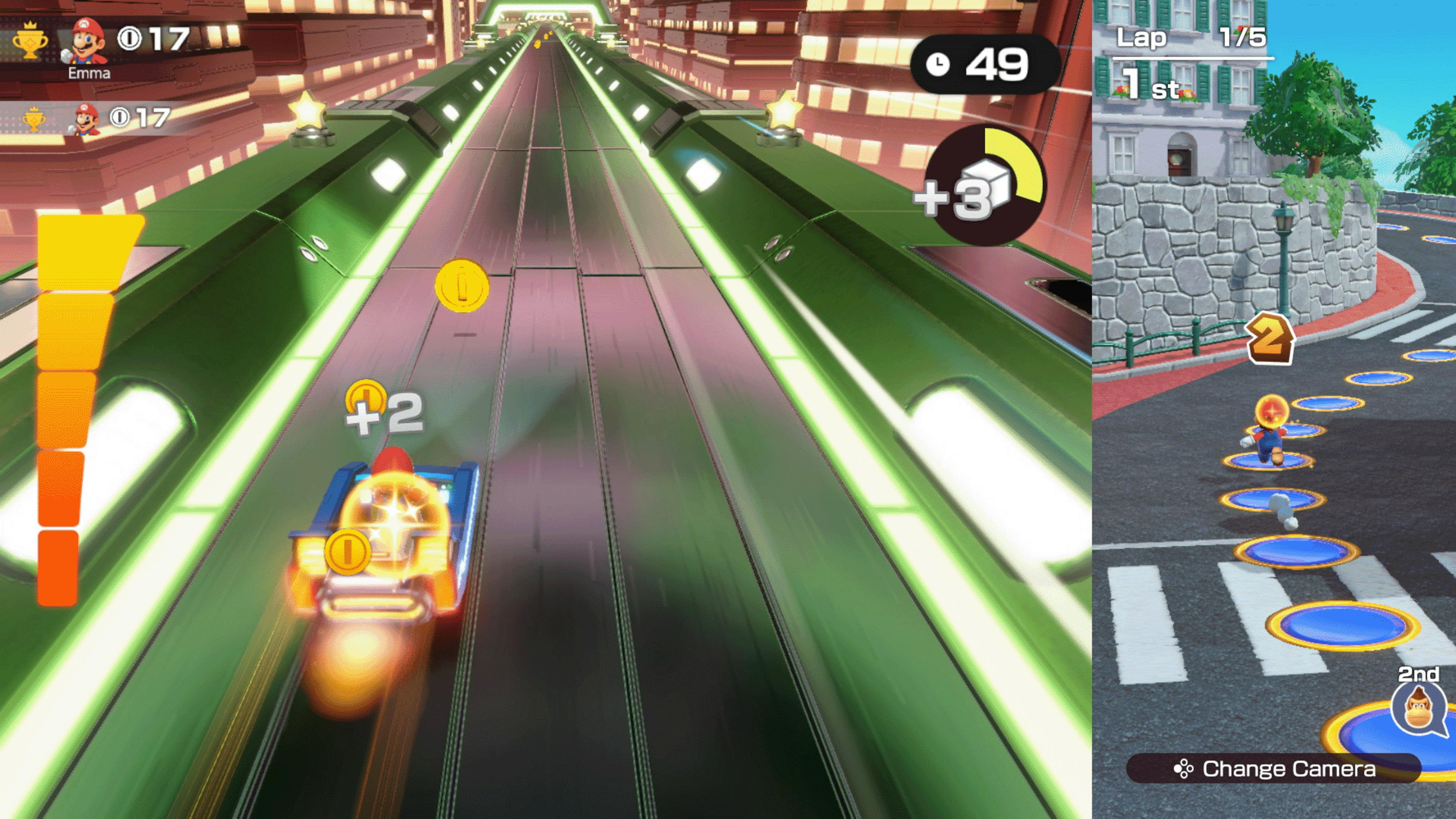 Ekran gry Mario Kart. Niebieski samochód ściga się na torze. Krajobraz miejski z budynkami i widokiem z boku ulicy.