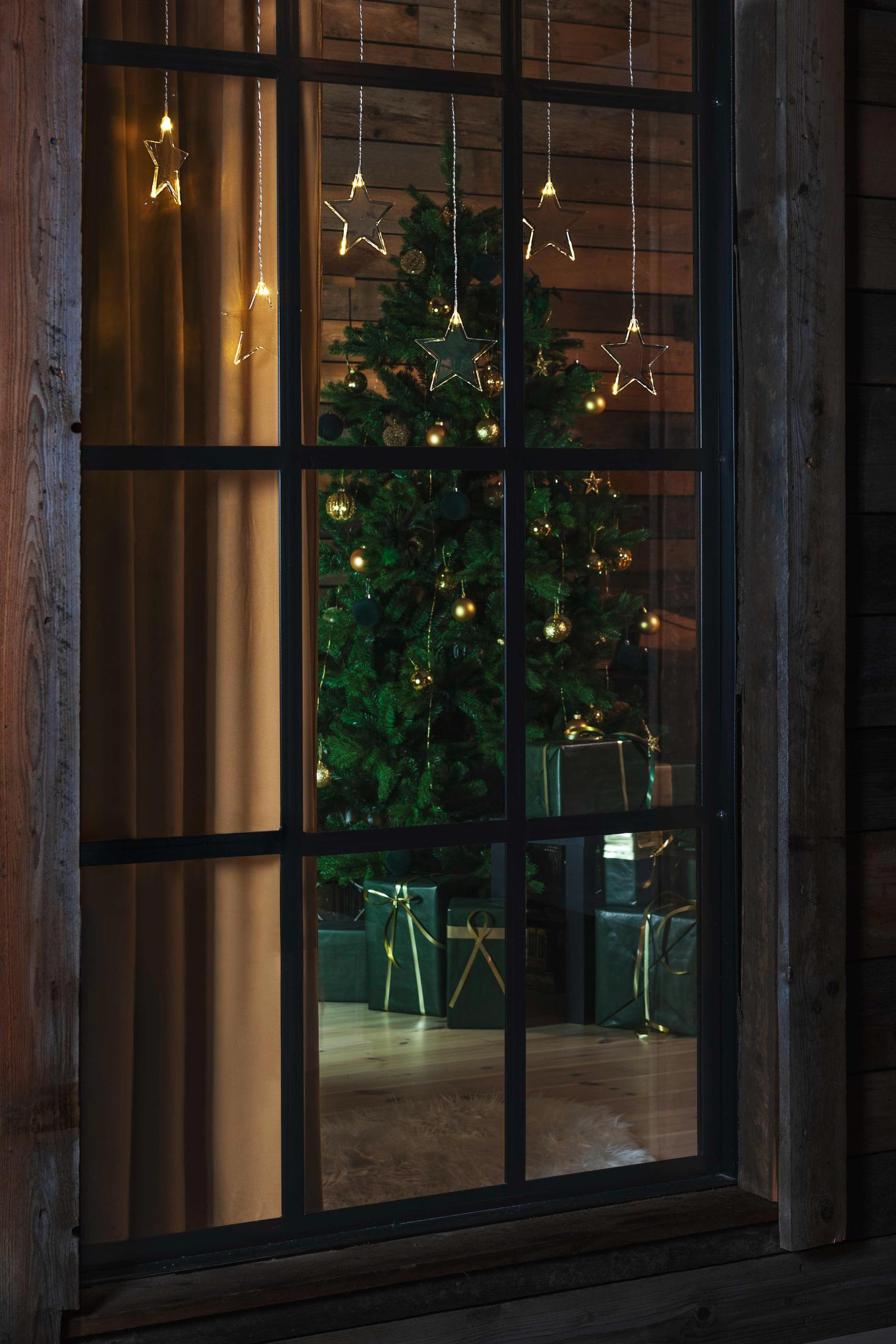 Blick durch ein Fenster auf einen geschmückten Weihnachtsbaum und Geschenke, mit Sternlichtern.