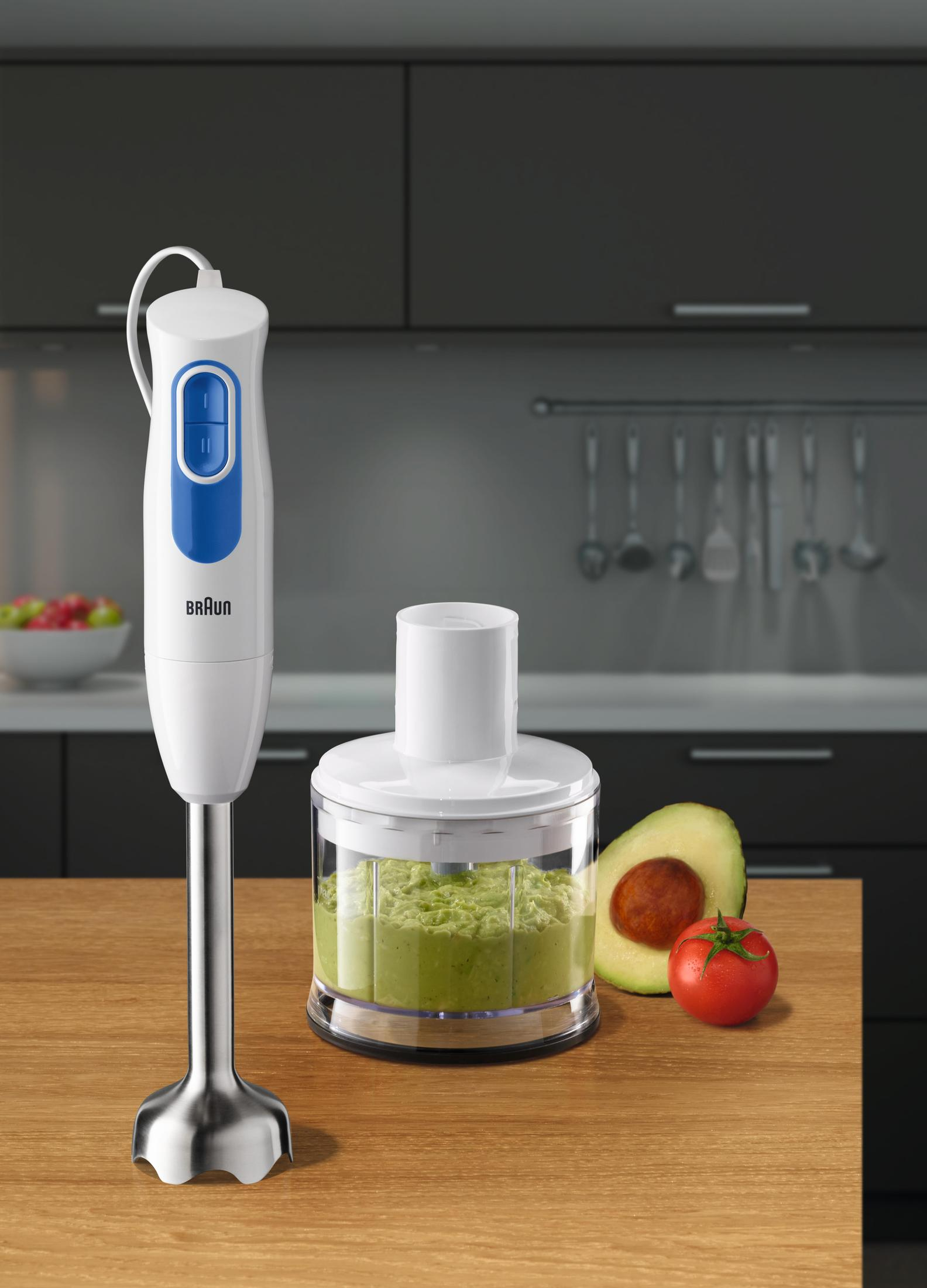 Biały blender ręczny z niebieskim przyciskiem, obok awokado, pomidora i posiekanej zieleni.