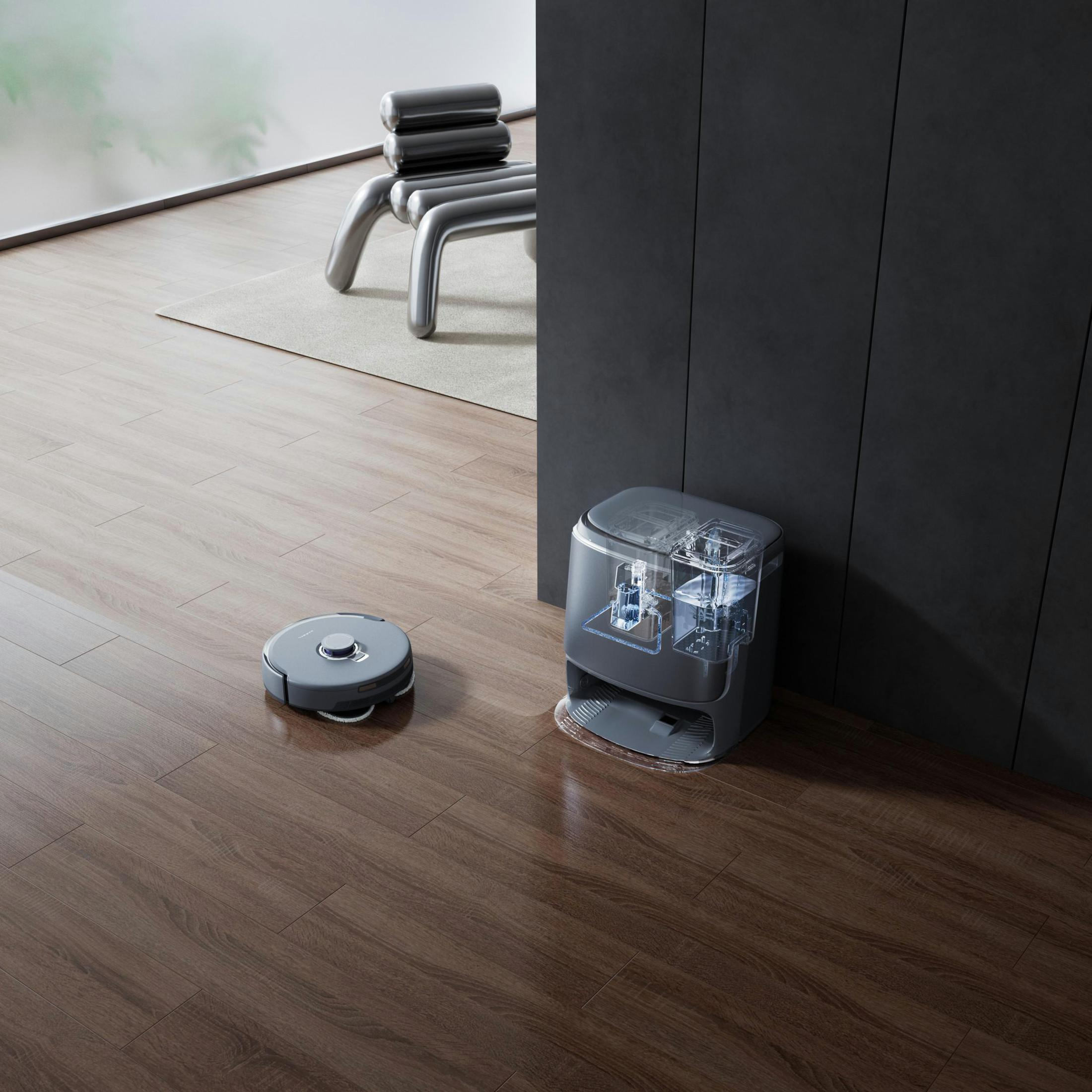 Ein Roboter-Staubsauger und seine Ladestation auf einem Holzboden. Im Hintergrund steht ein Stuhl. Die Station ist transparent.