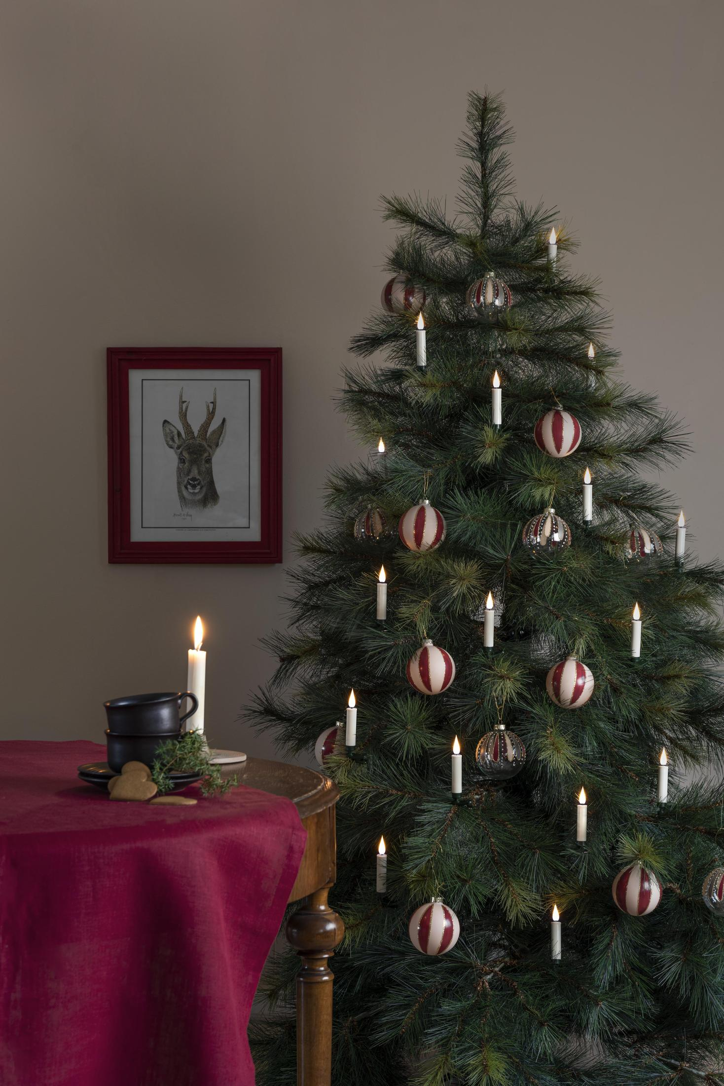Ein dekorierter Weihnachtsbaum neben einem Tisch mit Kerzen, einem Bild und einer Tasse. Der Raum ist schwach beleuchtet.