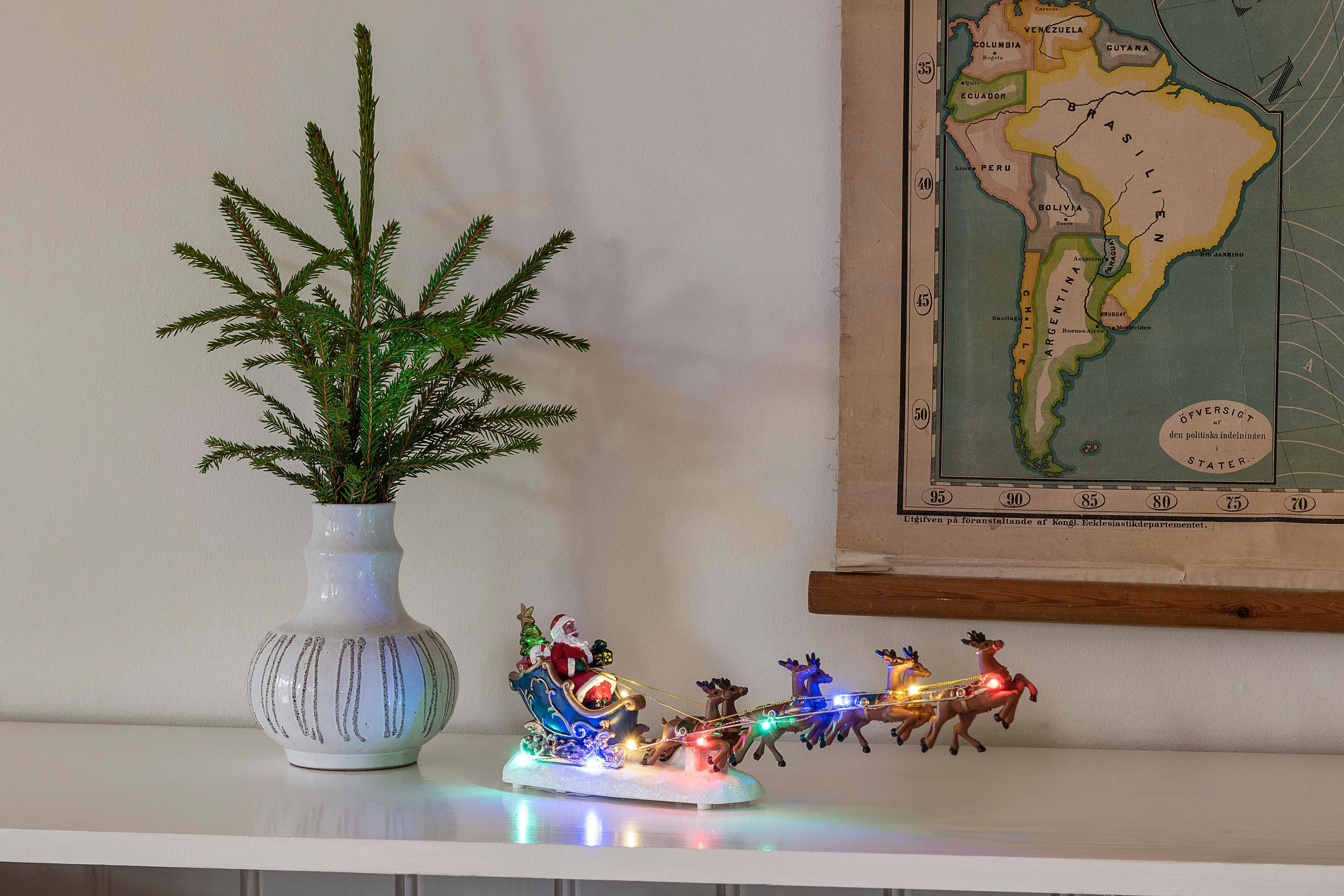Weihnachtsdeko: Eine weiße Vase mit Tanne und ein Santa-Schlitten mit Rentieren auf einem weißen Regal.