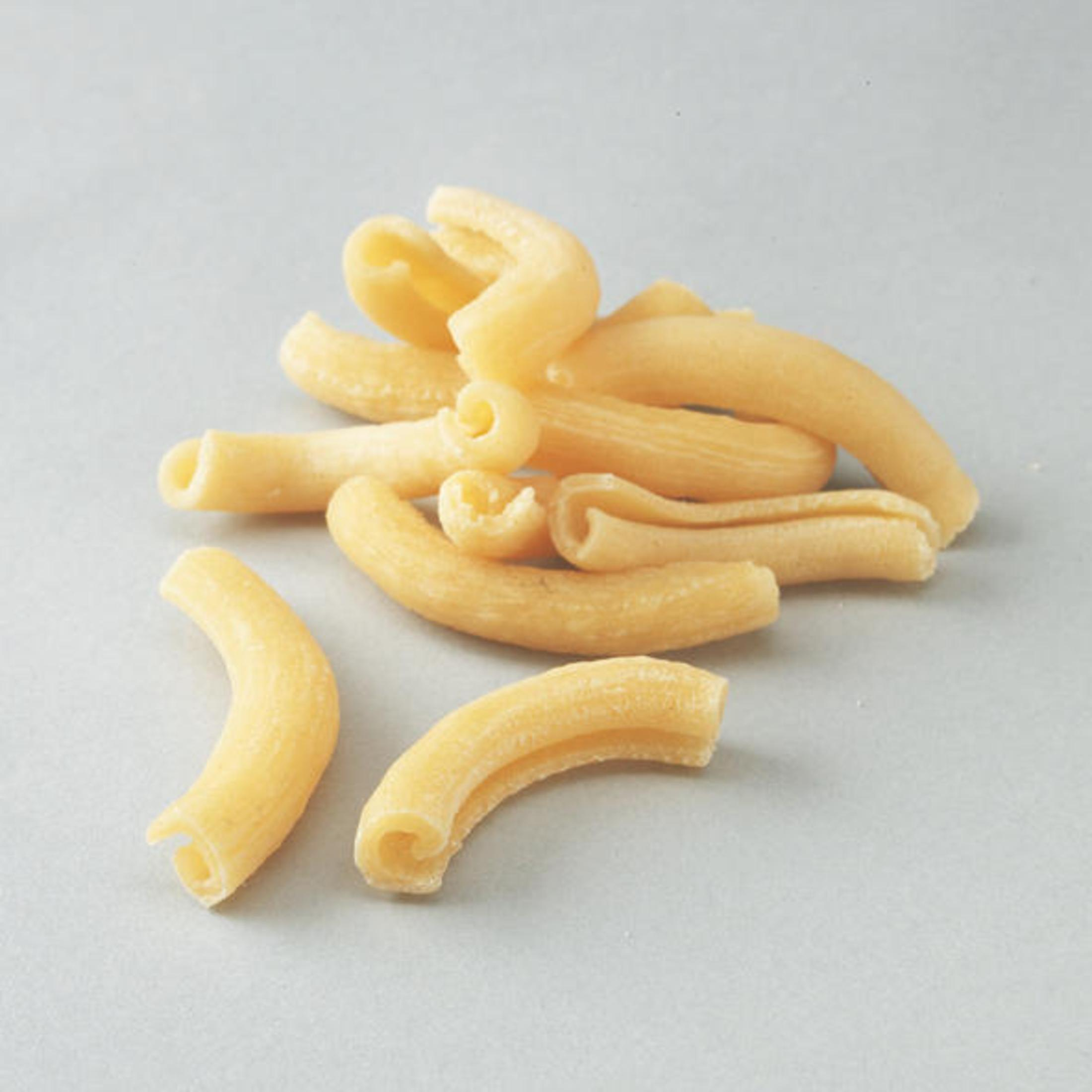 Pila di pasta di maccheroni a gomito su una superficie grigio chiaro. La pasta è di colore giallastro.