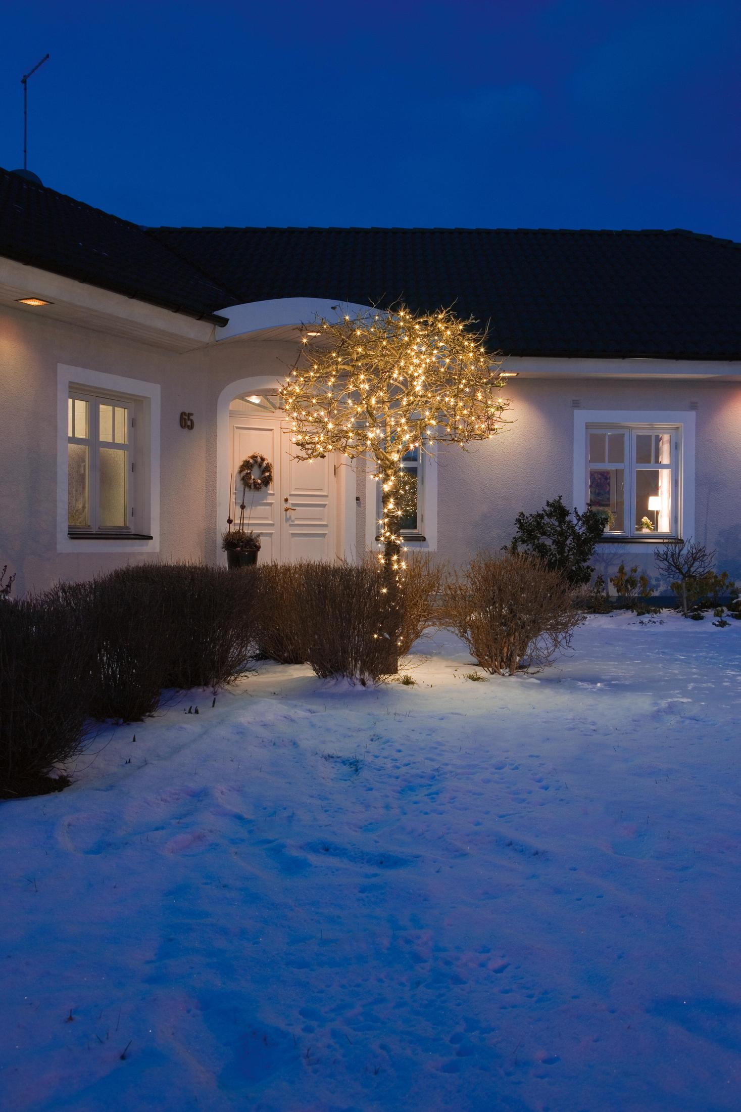 Una casa di notte con un cortile innevato e un albero decorato con luci.