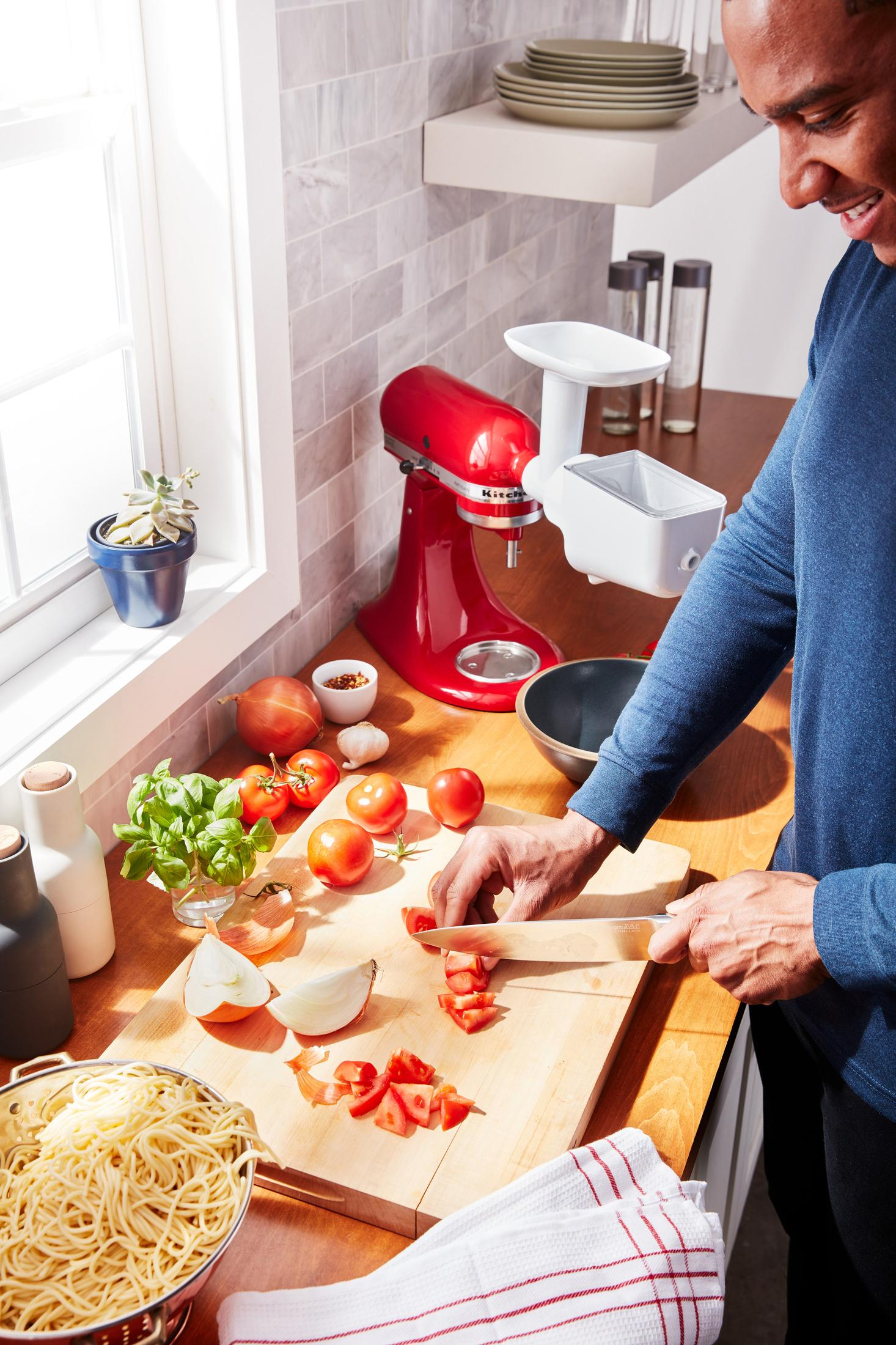 Una persona taglia i pomodori su un tagliere di legno vicino a un robot da cucina rosso e altri ingredienti.