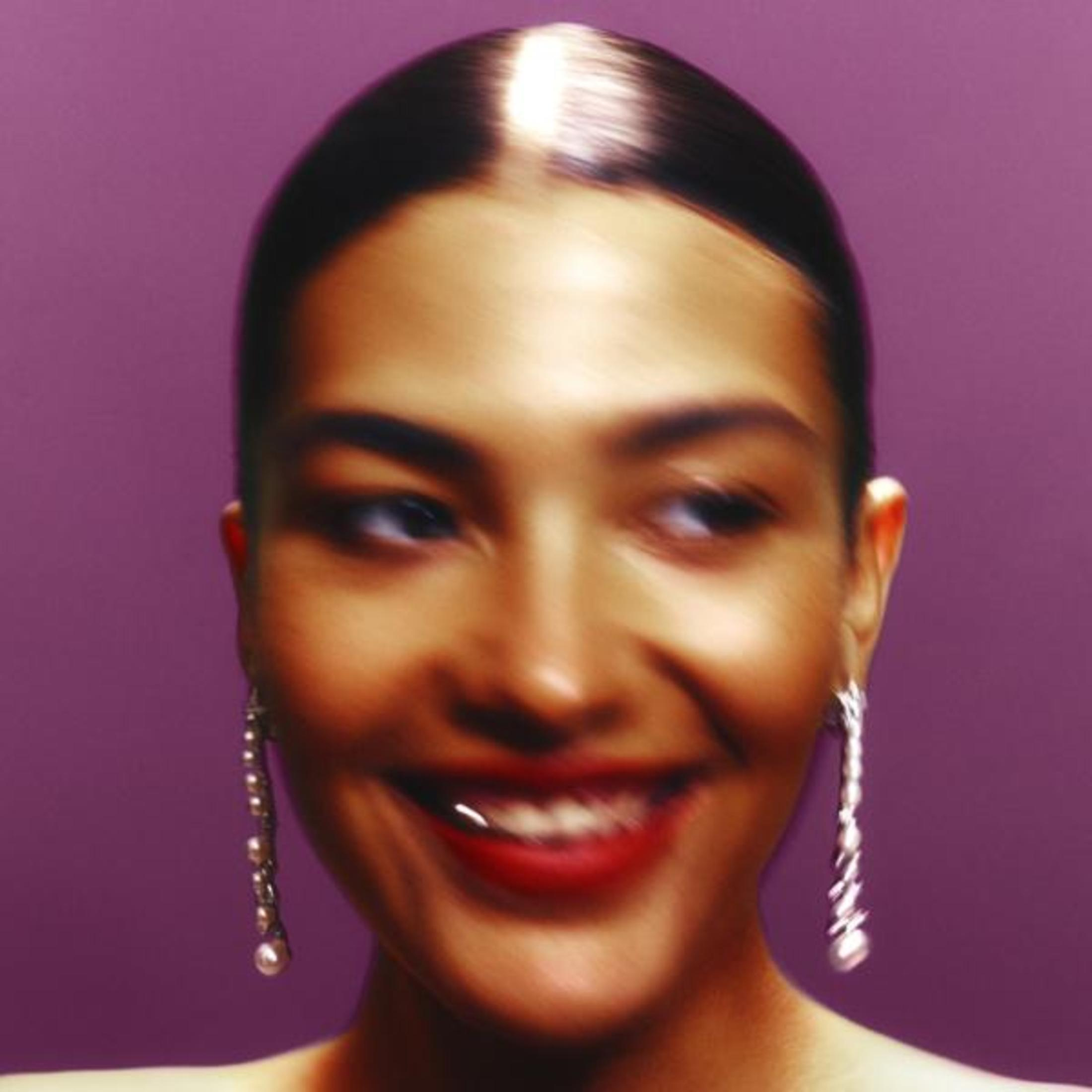 Il volto di una donna sorridente, con un leggero effetto sfocato. Indossa orecchini e ha una striscia bianca nei capelli.