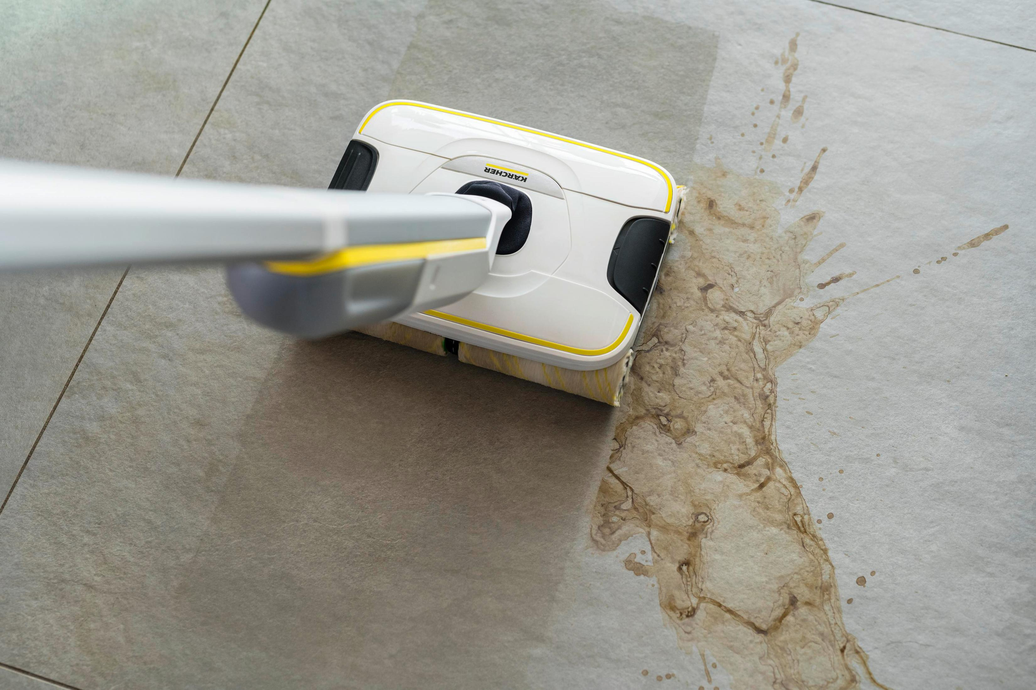 Una pulitrice per pavimenti bianca e gialla pulisce una macchia marrone su un pavimento piastrellato grigio chiaro.