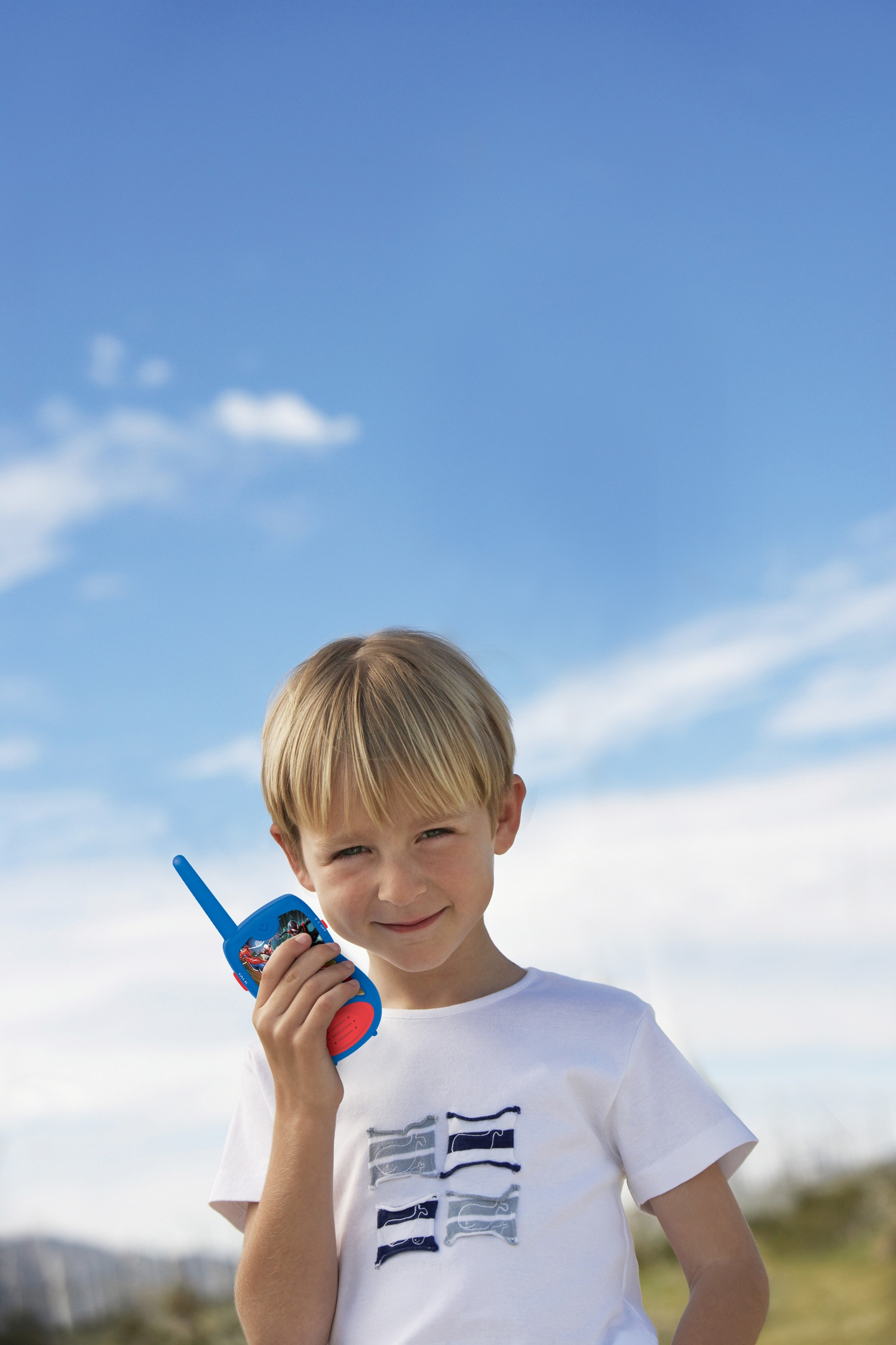 Niño con un walkie-talkie de Spider-Man, sonriendo. Cielo azul. Camisa blanca.