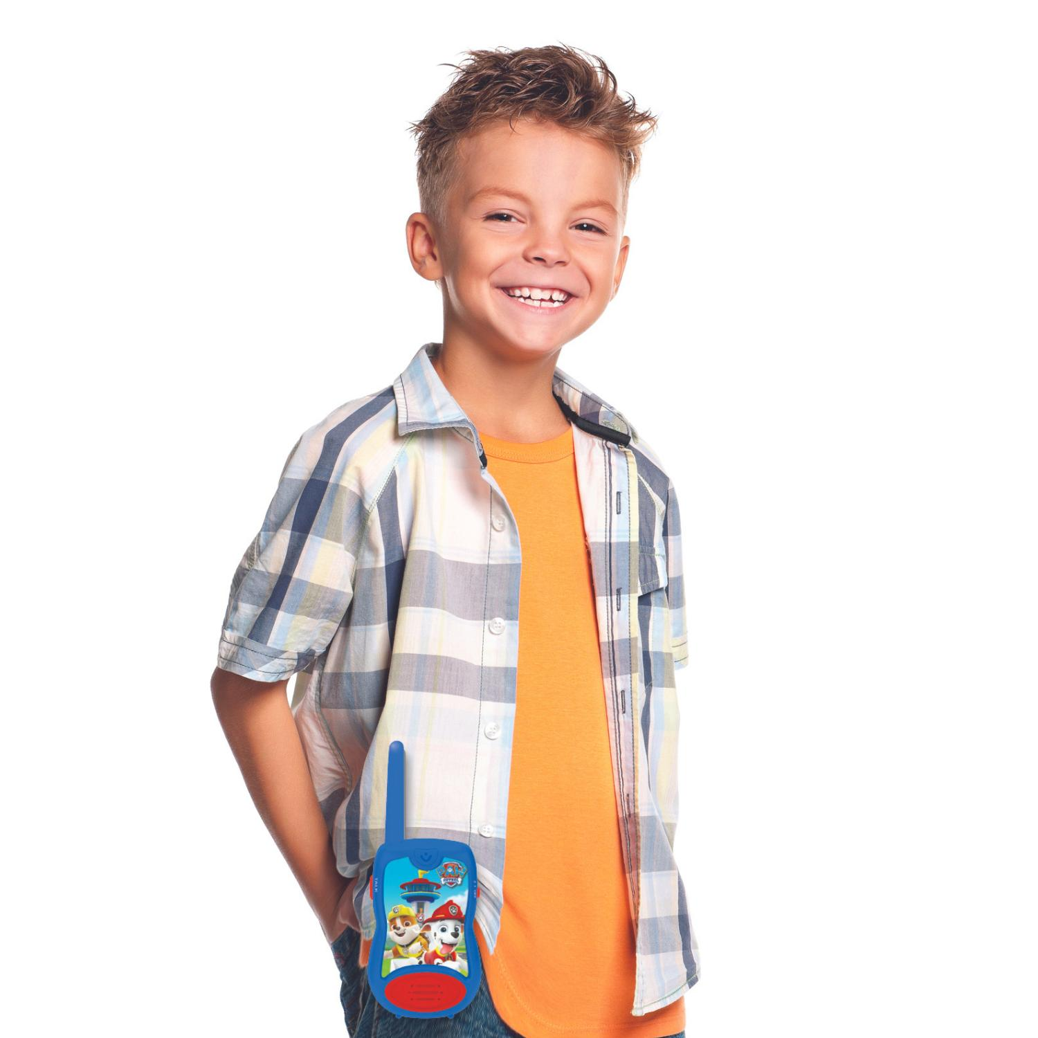 Uśmiechnięty chłopiec trzymający niebieskie walkie-talkie PAW Patrol, w koszuli w kratę i pomarańczowej koszulce.