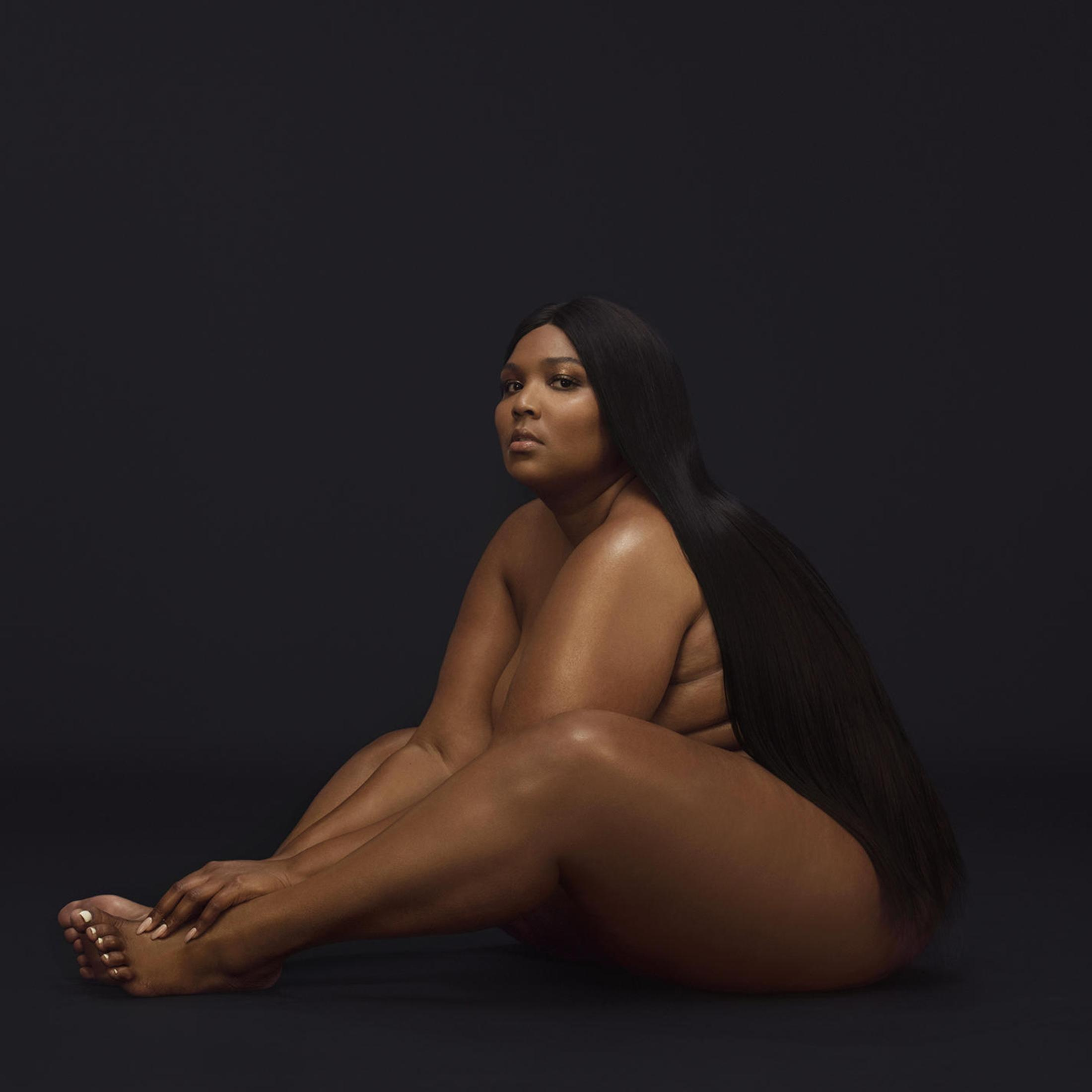 Lizzo, nue, assise avec de longs cheveux noirs couvrant son corps, sur fond noir.