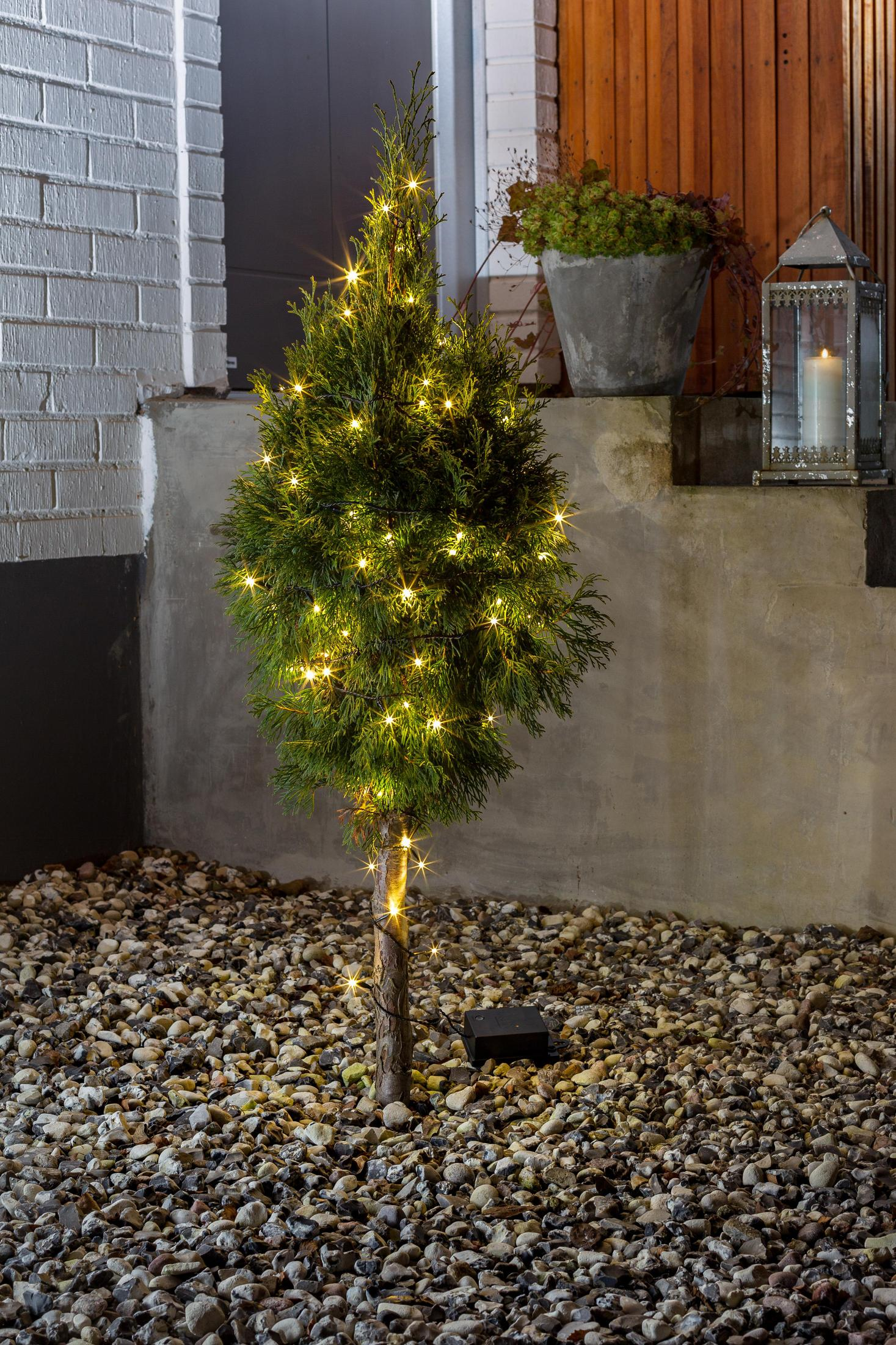Kleiner beleuchteter Baum mit Lichterketten, vor einer Wand und Steingrund.