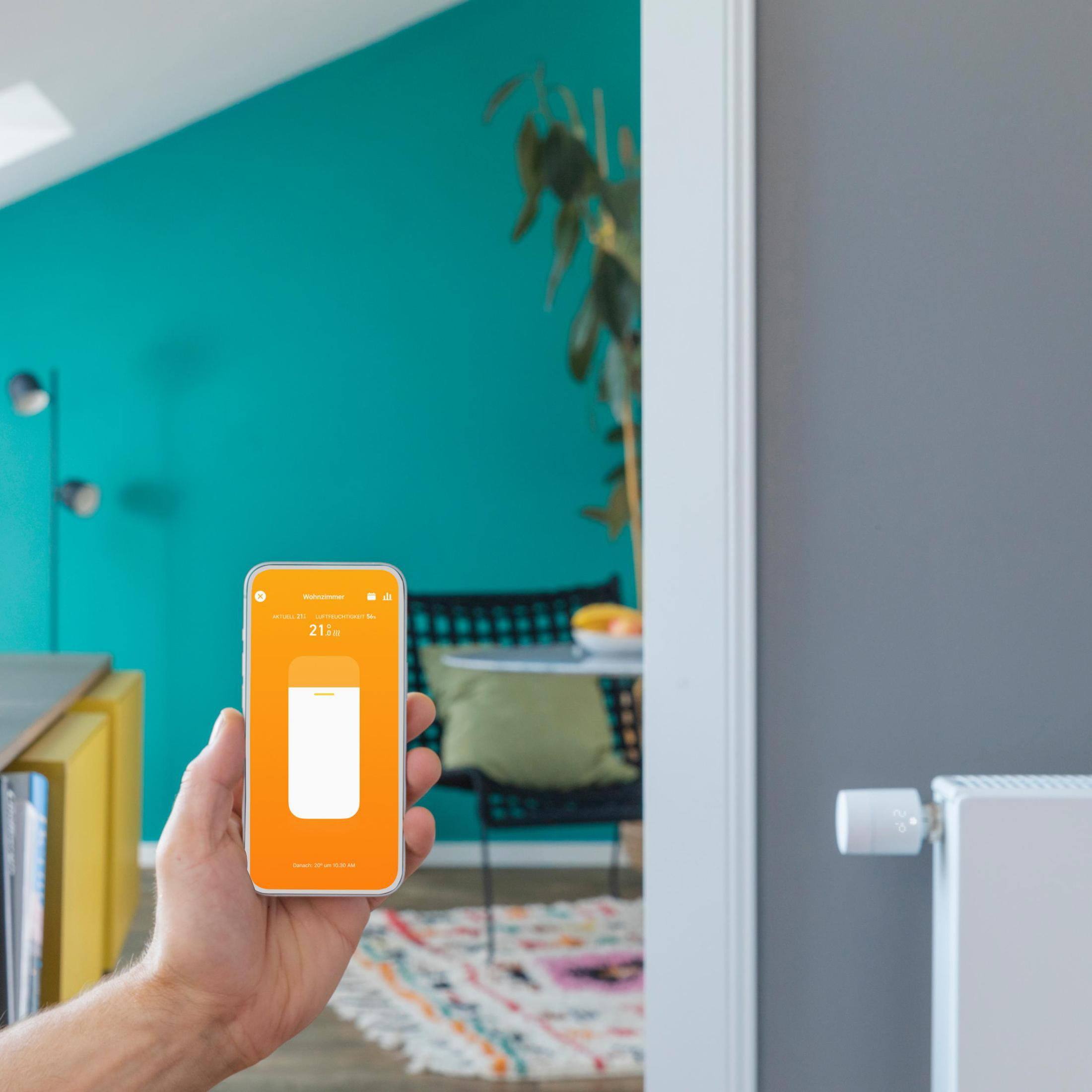 Hand, die ein Telefon hält, App zeigt Temperatur. Türkisfarbene Wand und Fensterrahmen.