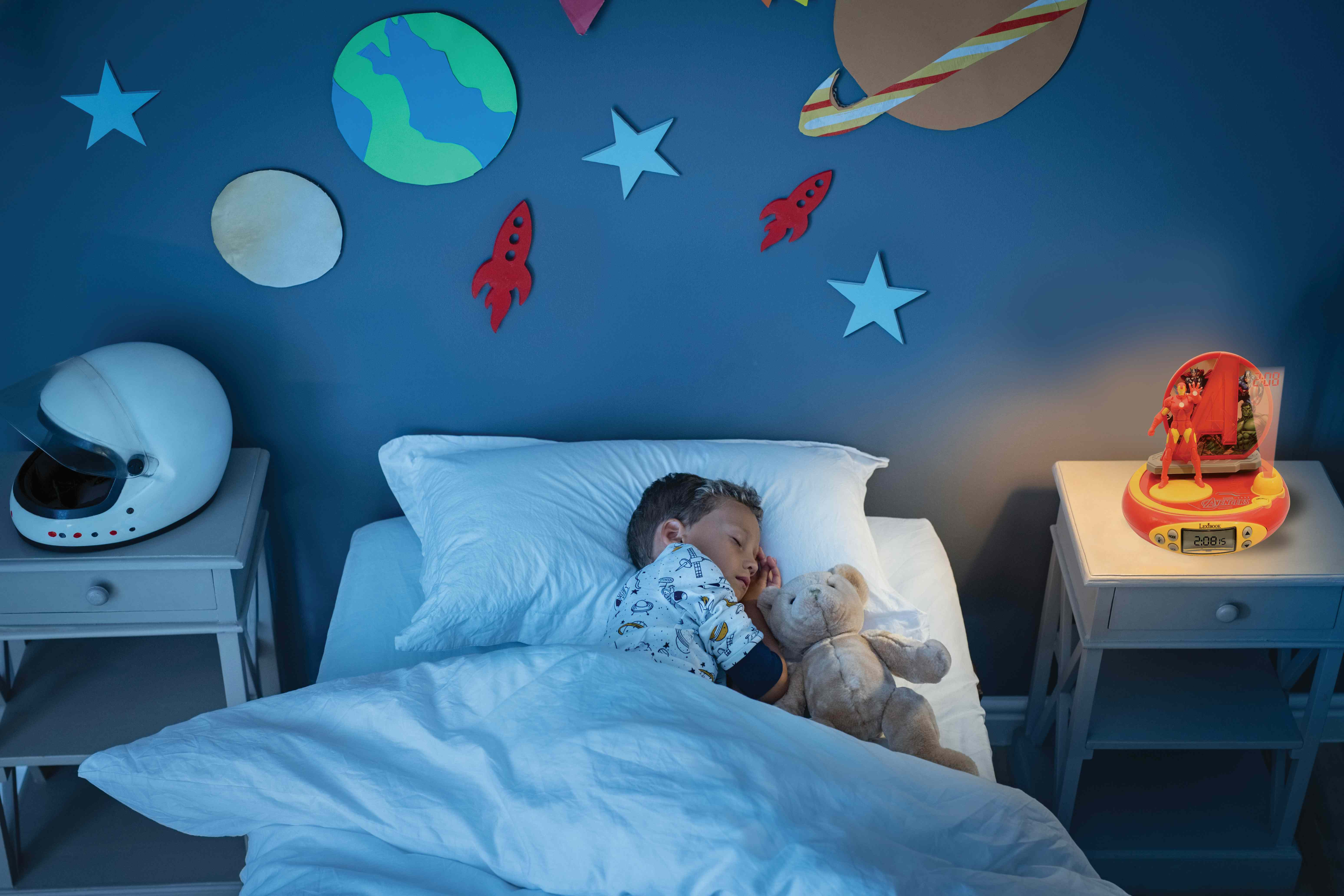 Un bambino dorme nel letto con un orsacchiotto, una luce notturna sul comodino.