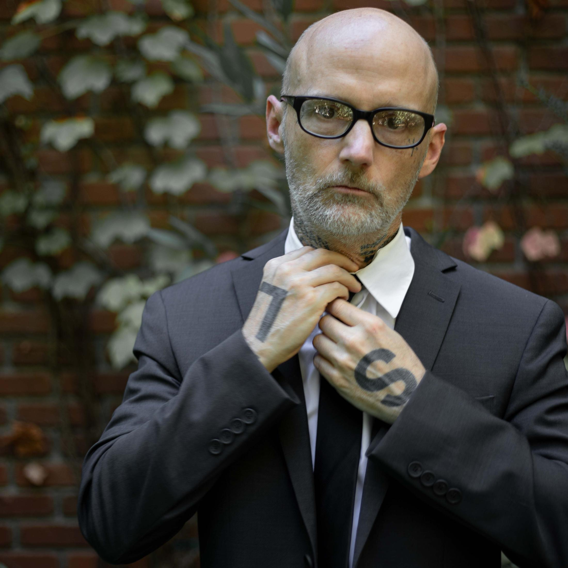 A man in a suit adjusts his tie. He has glasses and tattoos on his hands.