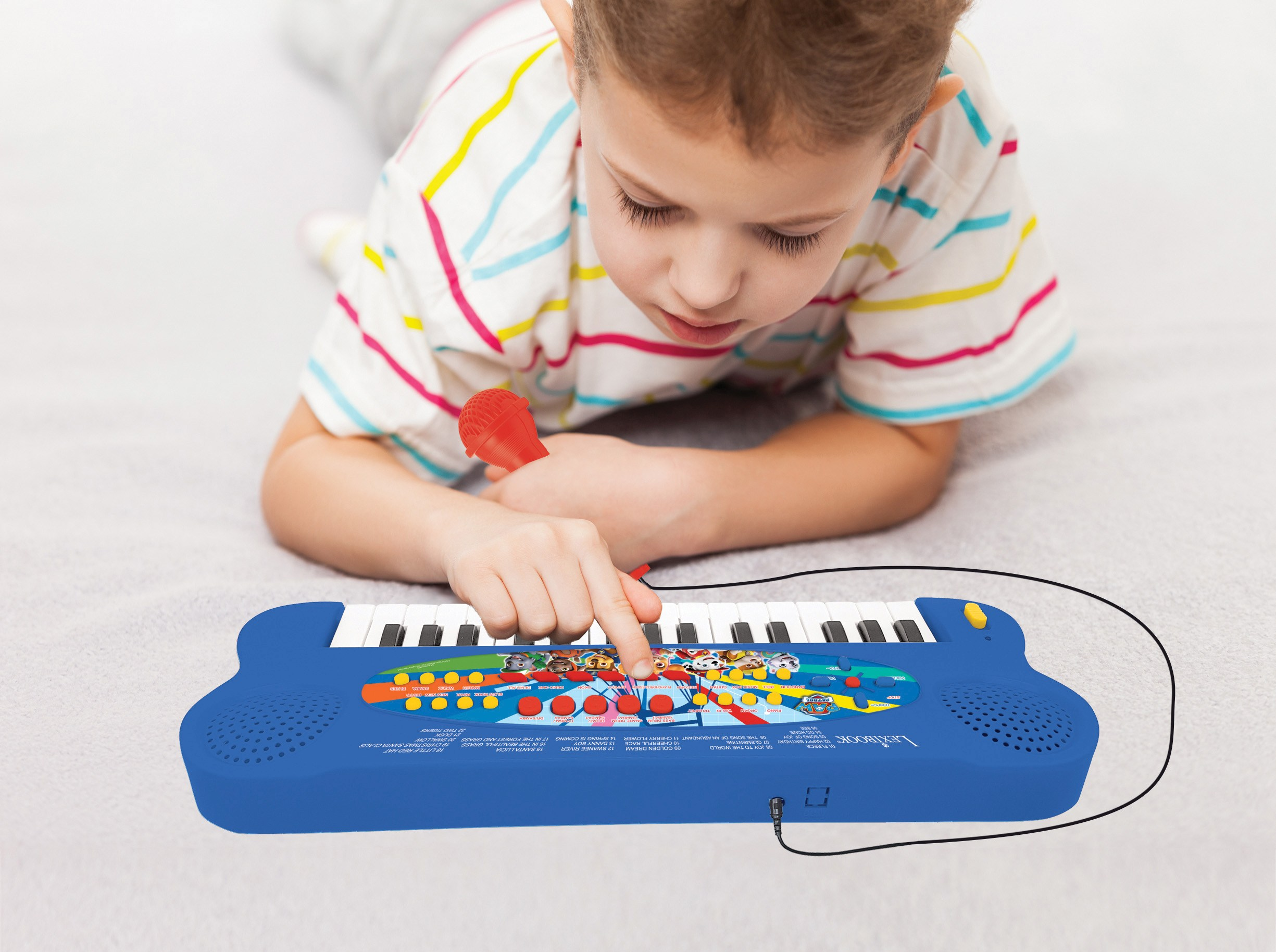 Ein Junge spielt ein blaues Spielzeugklavier mit Mikrofon. Das Klavier hat ein Paw Patrol-Thema, auf einem Bett.
