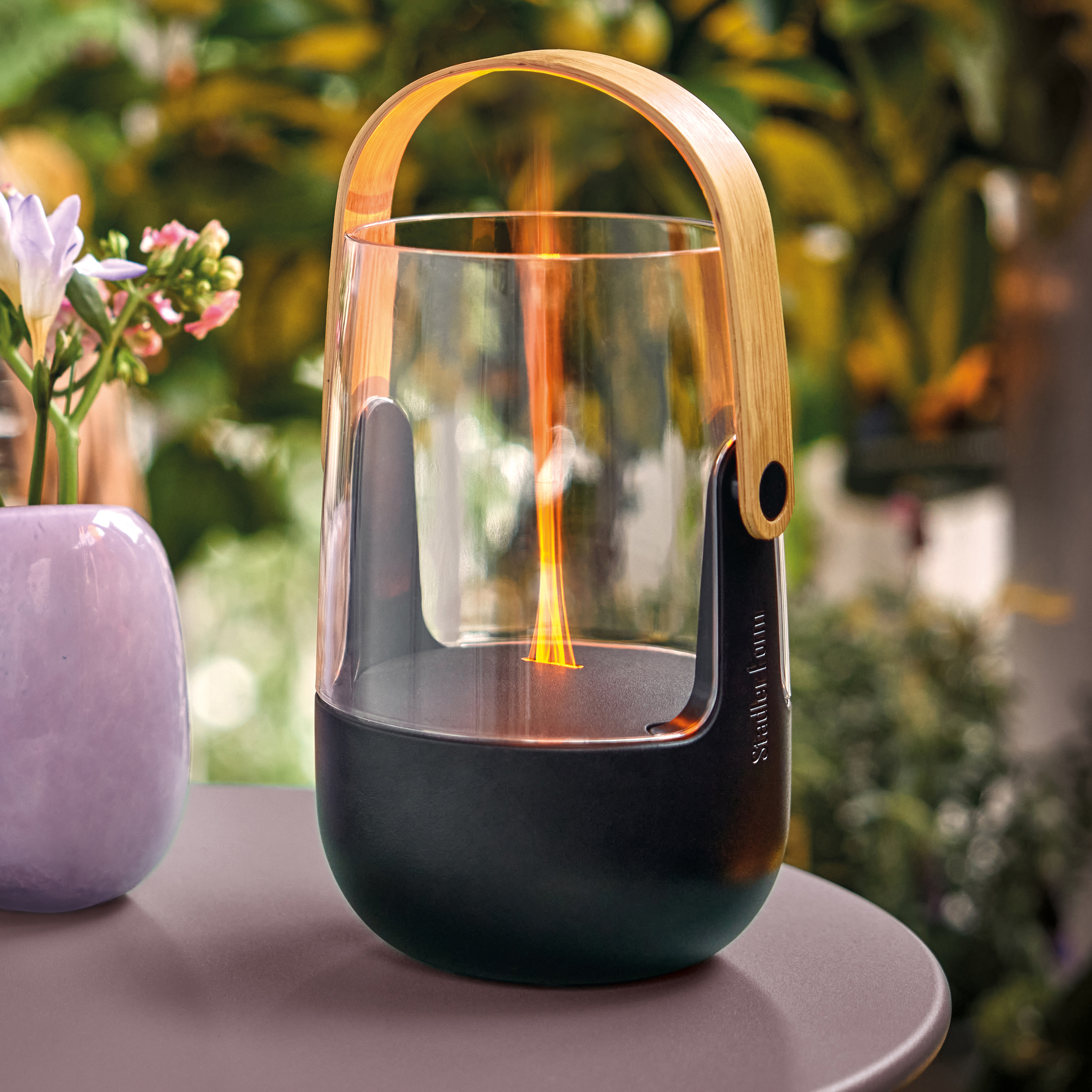 A decorative lantern with a flame, set outdoors on a table with plants.