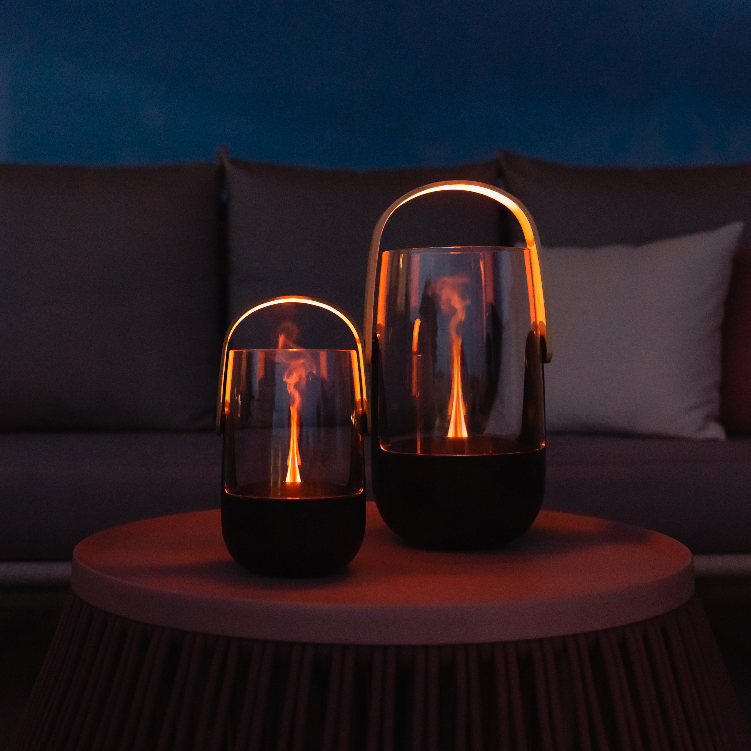 Two glass lanterns with flames on a table. A gray sofa is in the background.