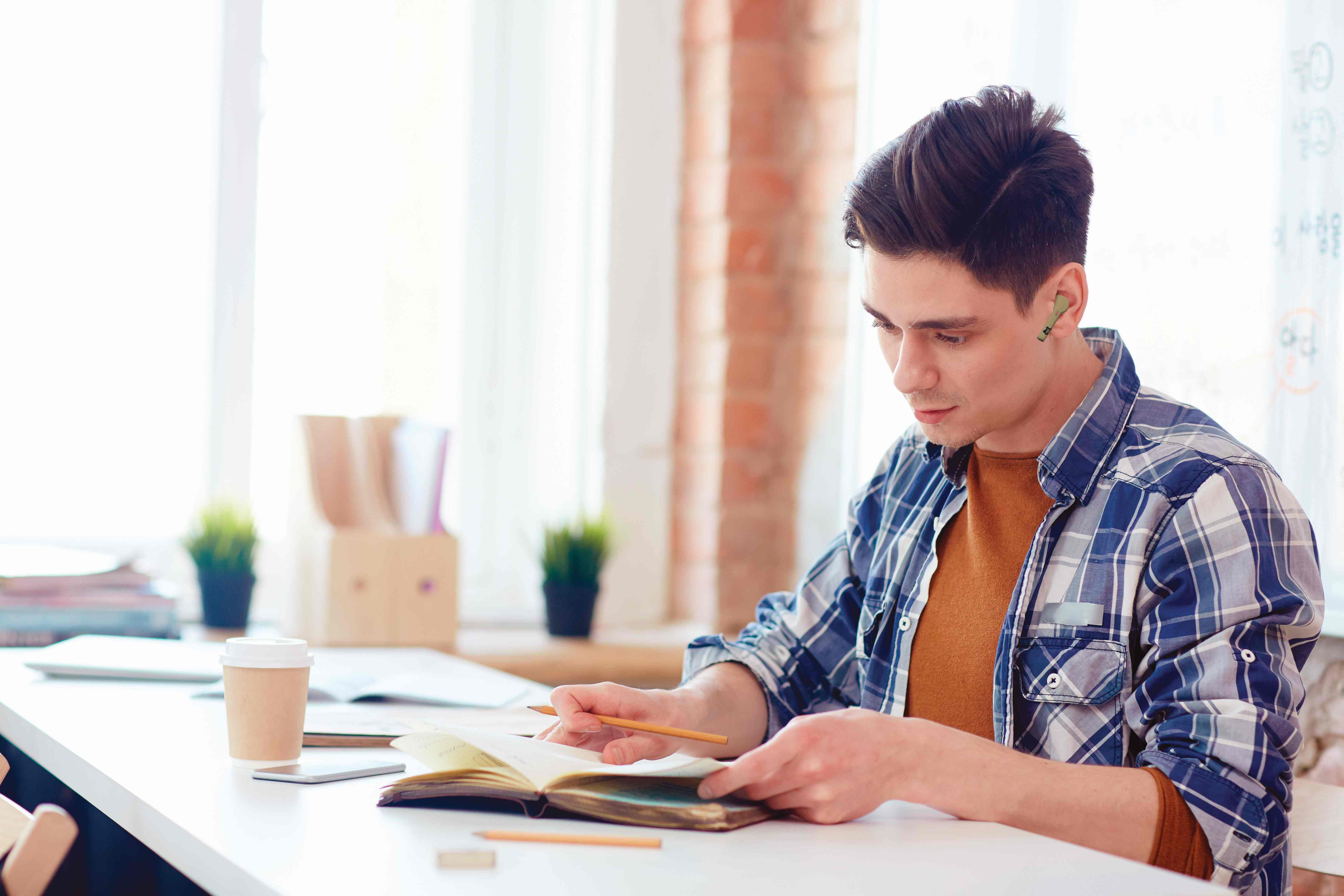 Uomo che usa gli auricolari mentre legge. Indossa una camicia a quadri. Libro e caffè sulla scrivania. Auricolari verdi.