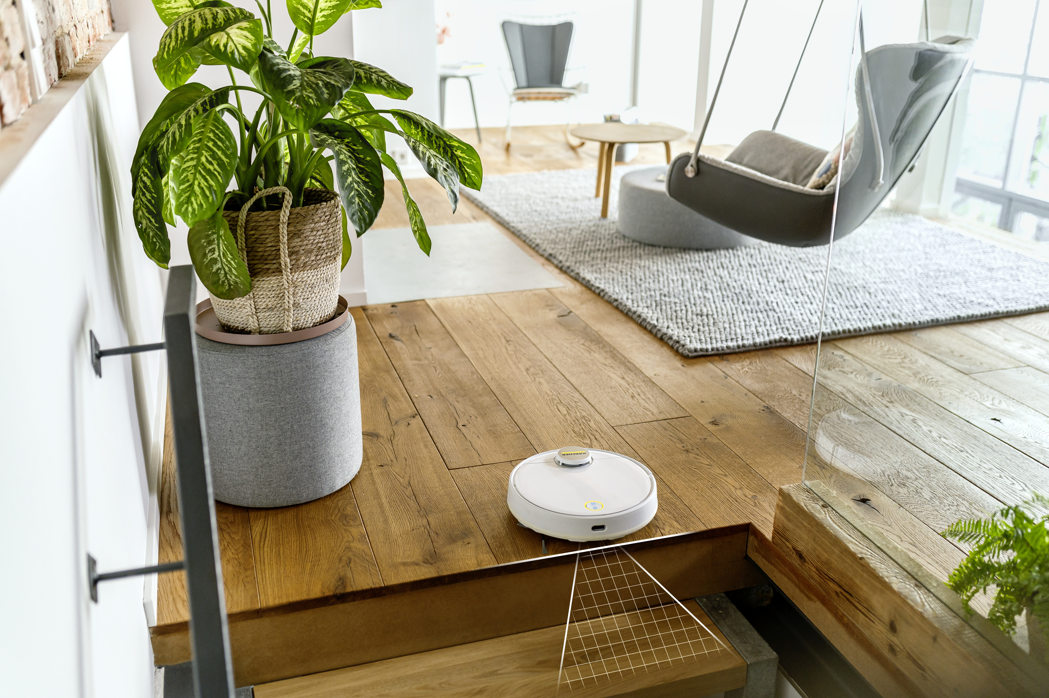 Una aspiradora robot blanca y dorada en un piso de madera. Se ven una planta y una silla colgante.