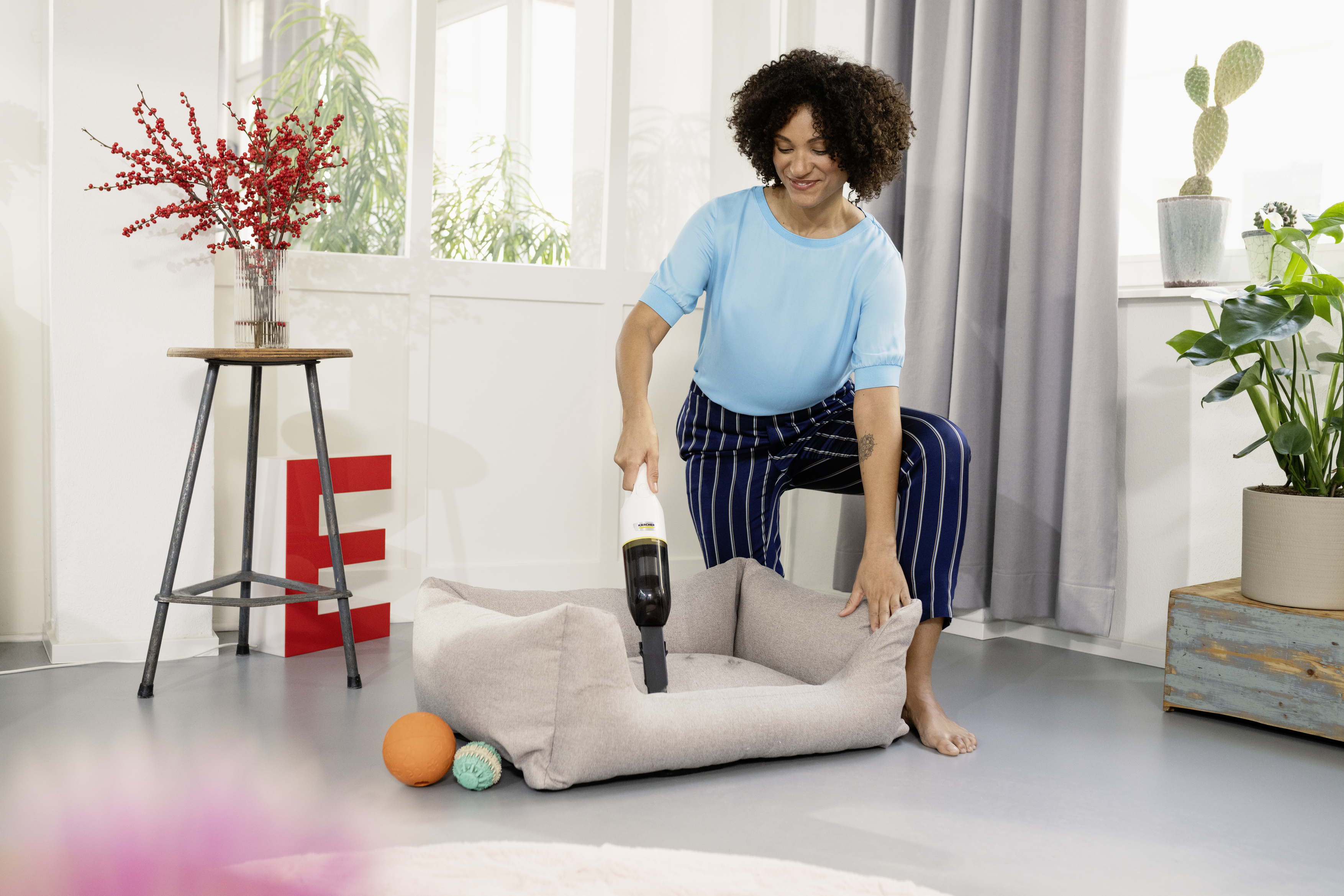 Een vrouw die een hondenmand stofzuigt in een kamer. Een vaas met bloemen en een rode 'E' zijn zichtbaar.
