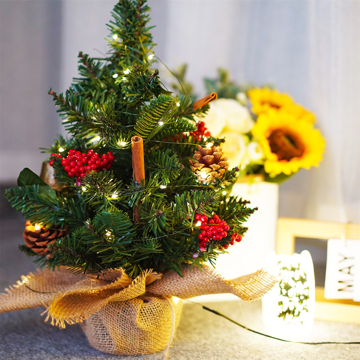 Ein kleiner Weihnachtsbaum mit Lichtern, Tannenzapfen und roten Beeren. Eine Vase mit Sonnenblumen steht dahinter.