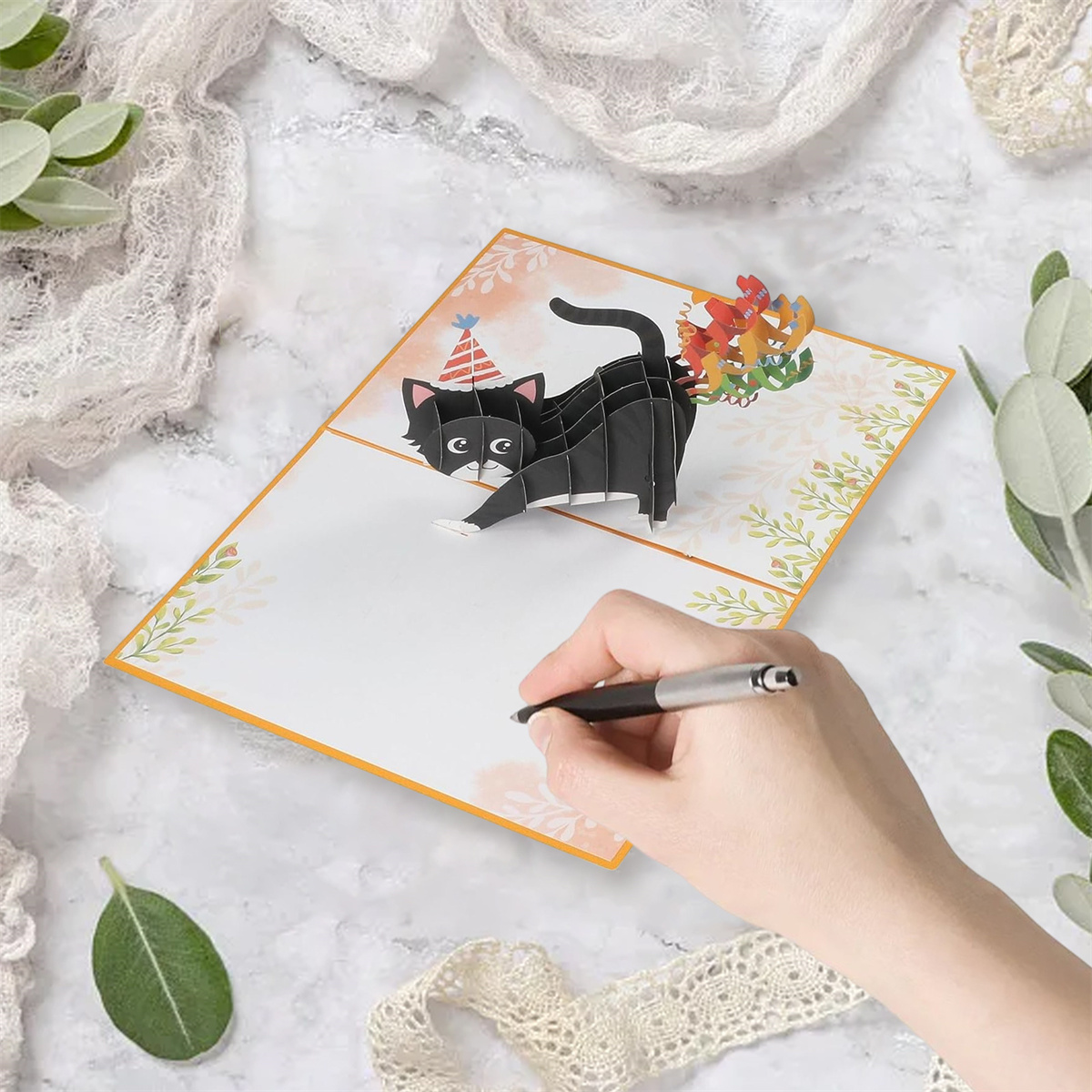 Eine Hand schreibt auf eine Pop-up-Karte mit einer schwarzen Katze. Der Hintergrund ist eine Marmoroberfläche.