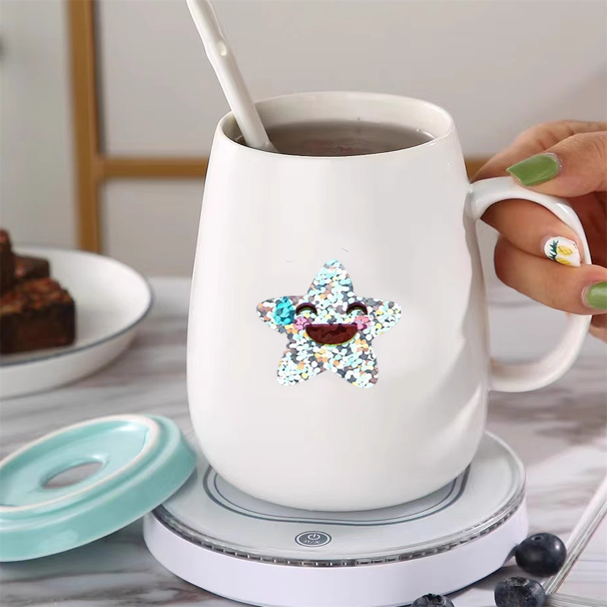 Eine weiße Tasse mit Blumenstern-Aufkleber, Löffel und Heizsockel stehen auf einem weißen Tisch.