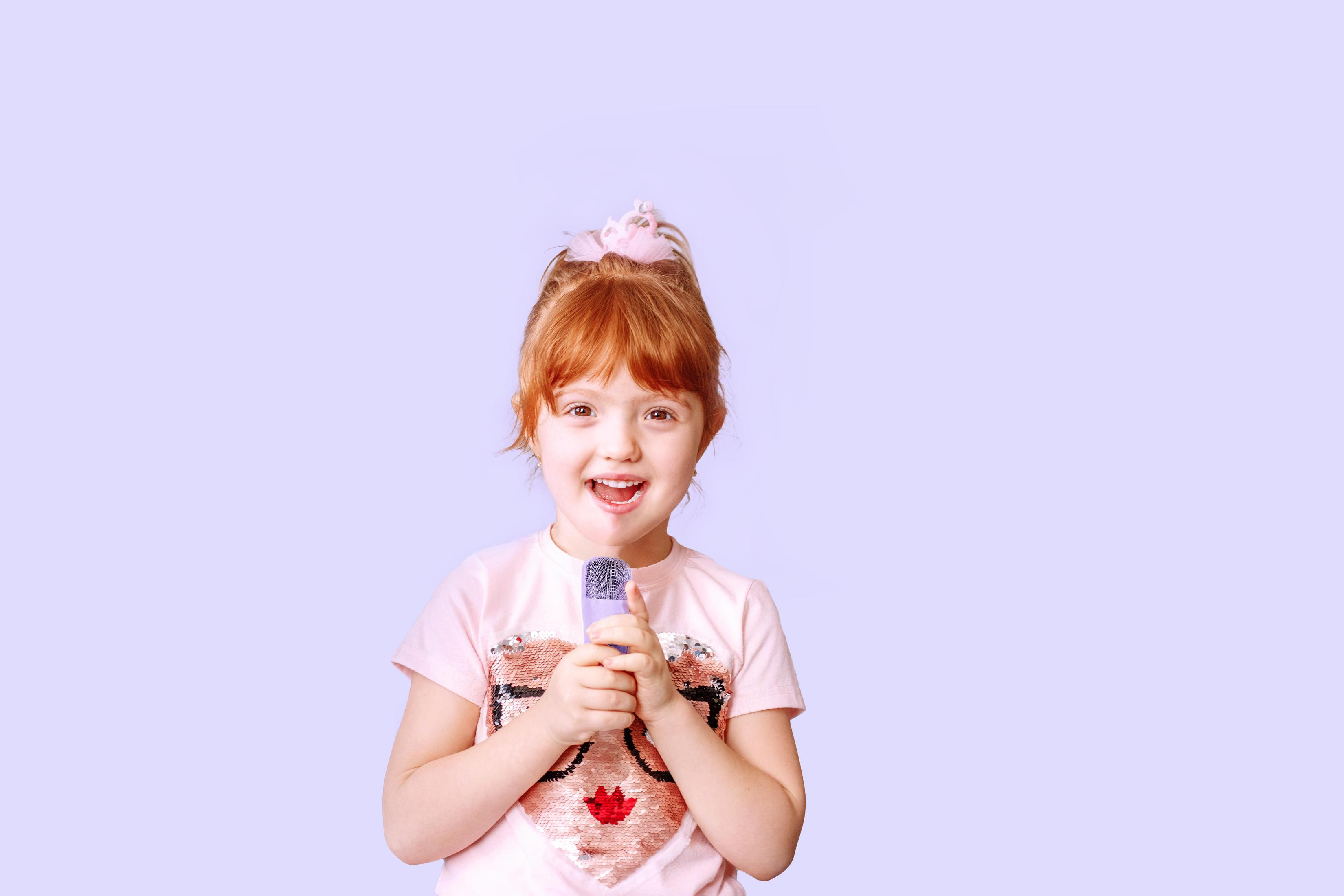 Une jeune fille aux cheveux roux chantant dans un microphone, souriant devant un fond violet clair.